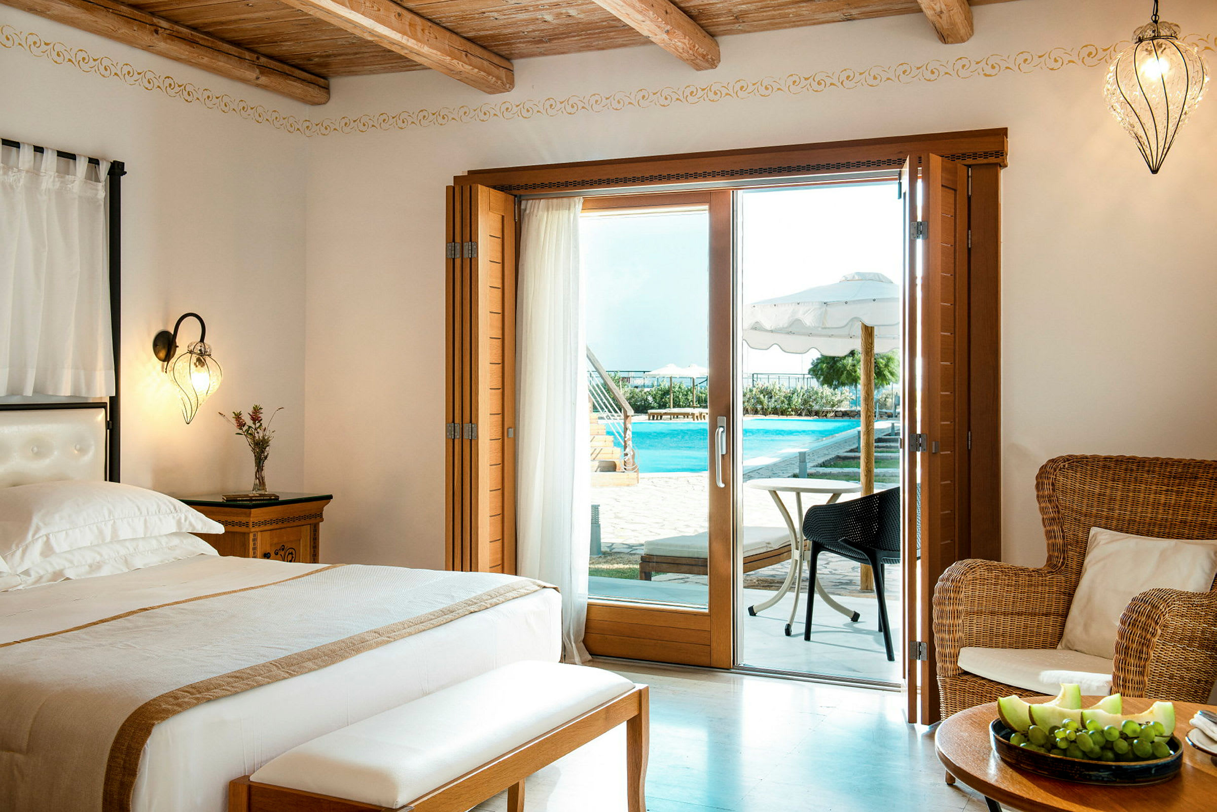 Ett hotellrum med en dubbelsäng i fokus, en öppen glasdörr med utsikt mot en pool och en fruktassiett på ett träbord. (AI-genererad text)