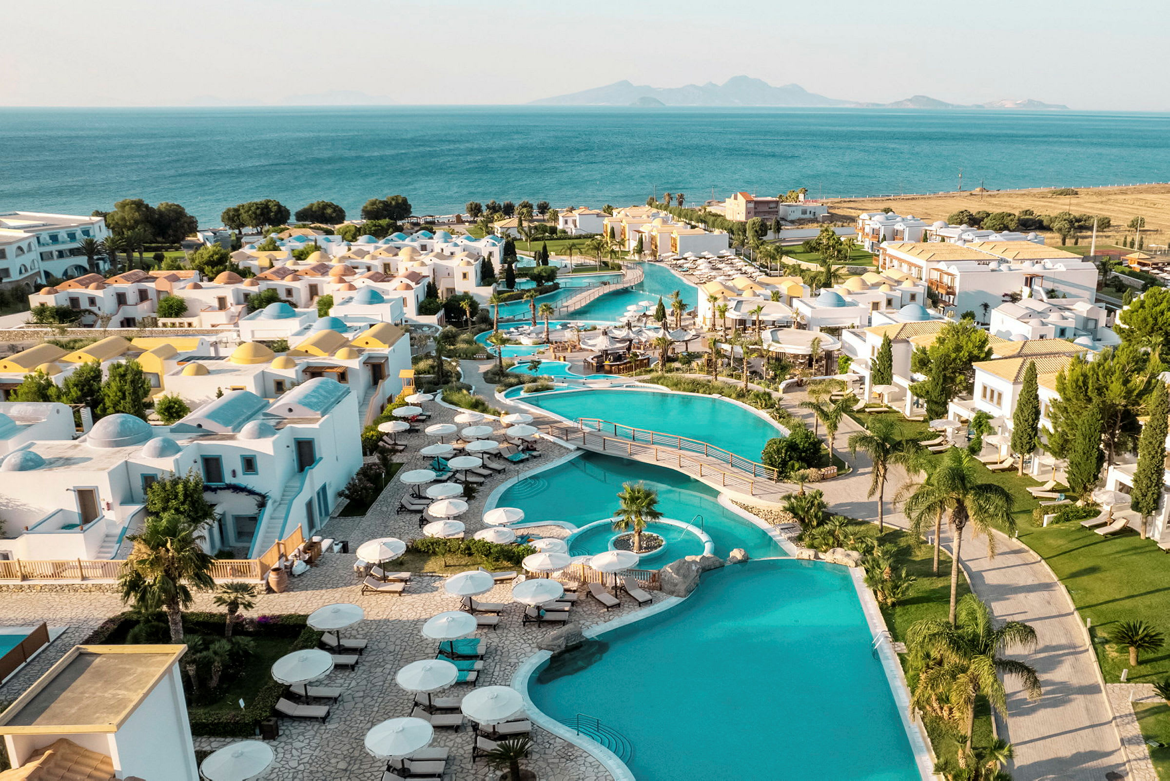 Vy över ett semesterkomplex med pooler, vita byggnader och utsikt mot havet i bakgrunden. (AI-genererad text)