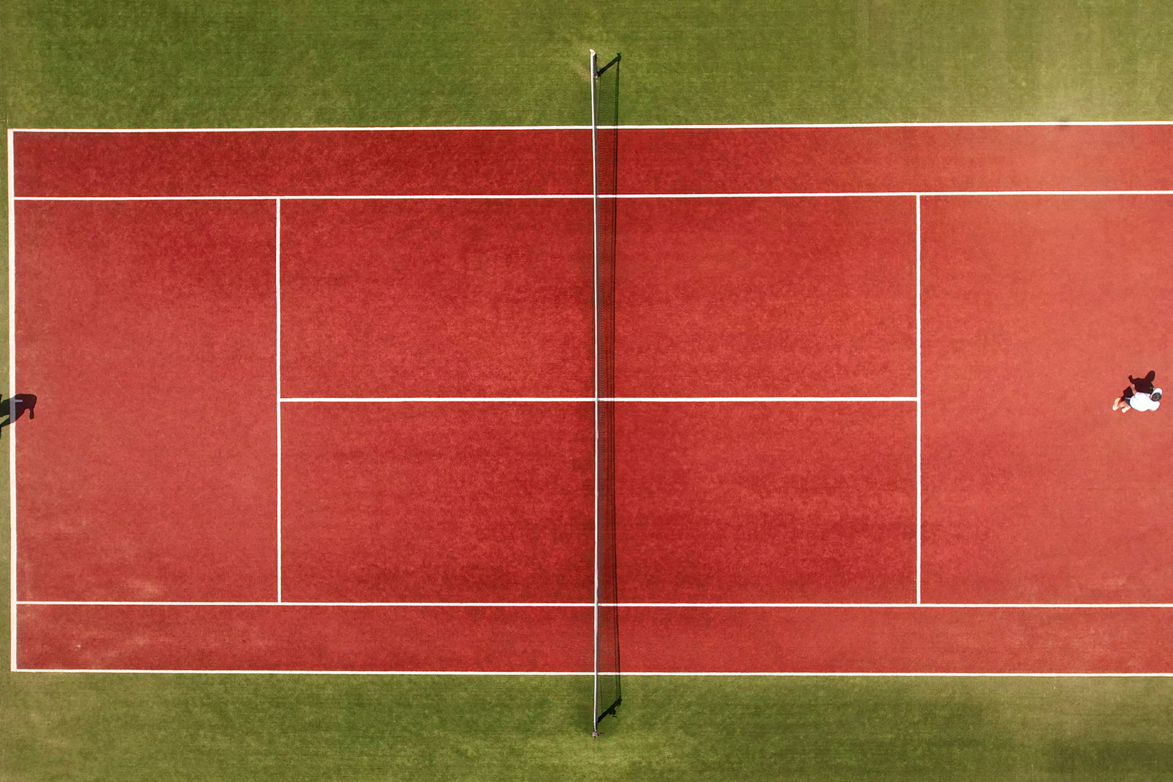 Flygbild av en röd tennisbana med nät, två personer syns spelande. (AI-genererad text)