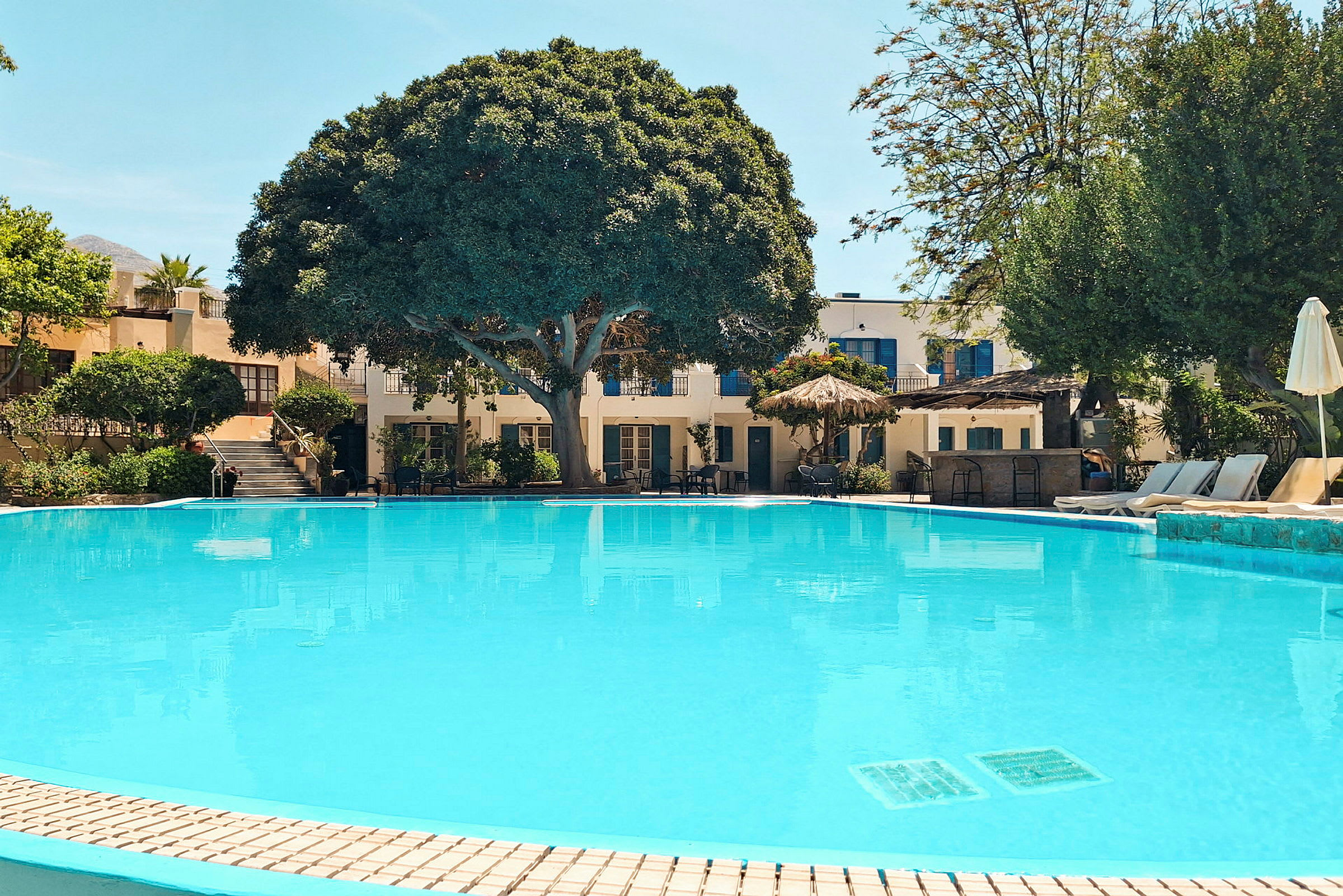 En stor swimmingpool framför ett hotell med omgivande grönska och parasoller. (AI-genererad text)