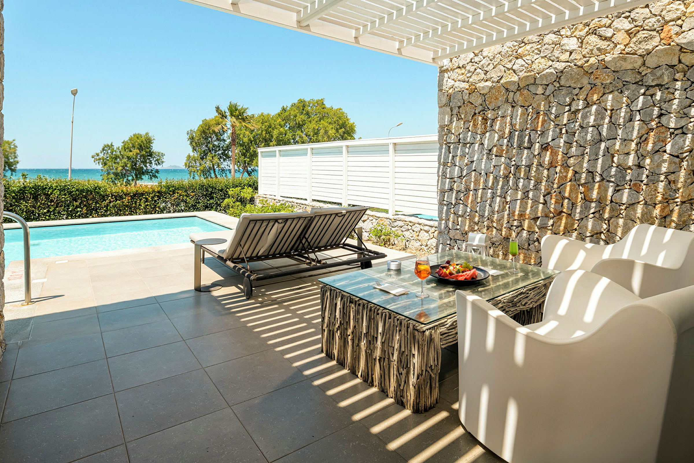 Terrass med utemöbler och solbädd vid en pool, med havsutsikt i bakgrunden. (AI-genererad text)