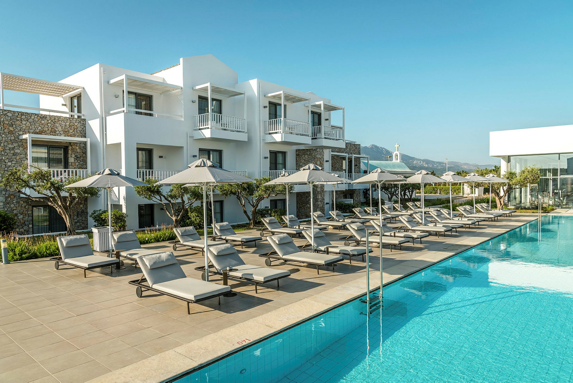 En hotellpool med solsängar och parasoller, omgiven av vita byggnader. (AI-genererad text)