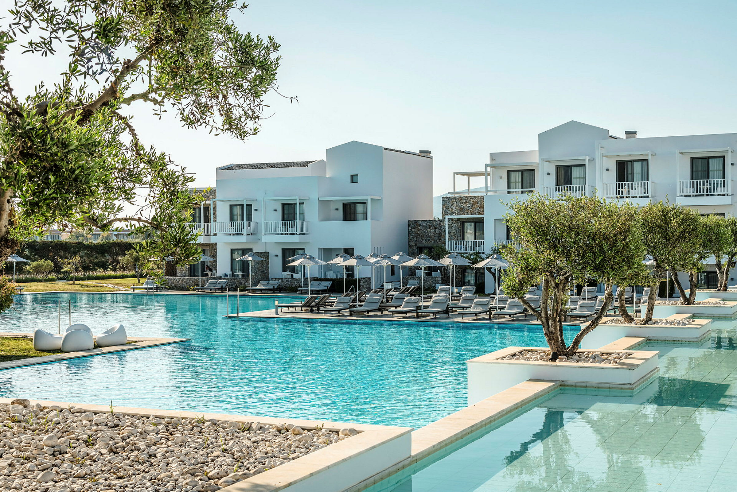 Bilden visar ett hotell med vit fasad vid en stor pool, omgiven av solstolar och parasoller samt grönska. (AI-genererad text)