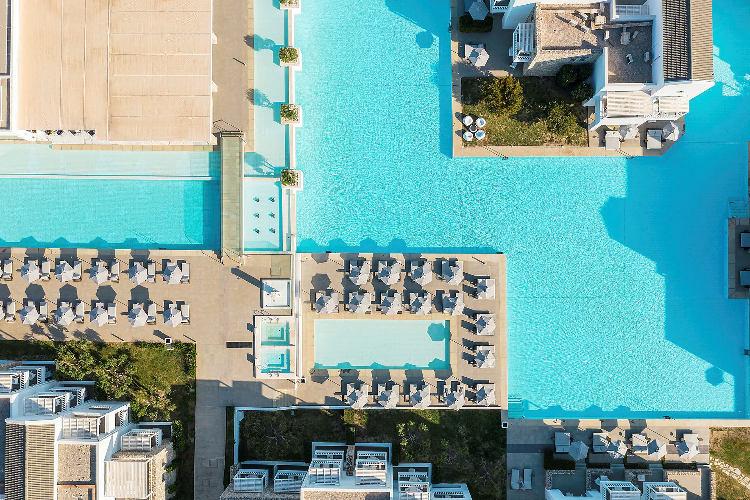 Flygfoto av en större poolanläggning med solstolar och byggnader omkring. (AI-genererad text)