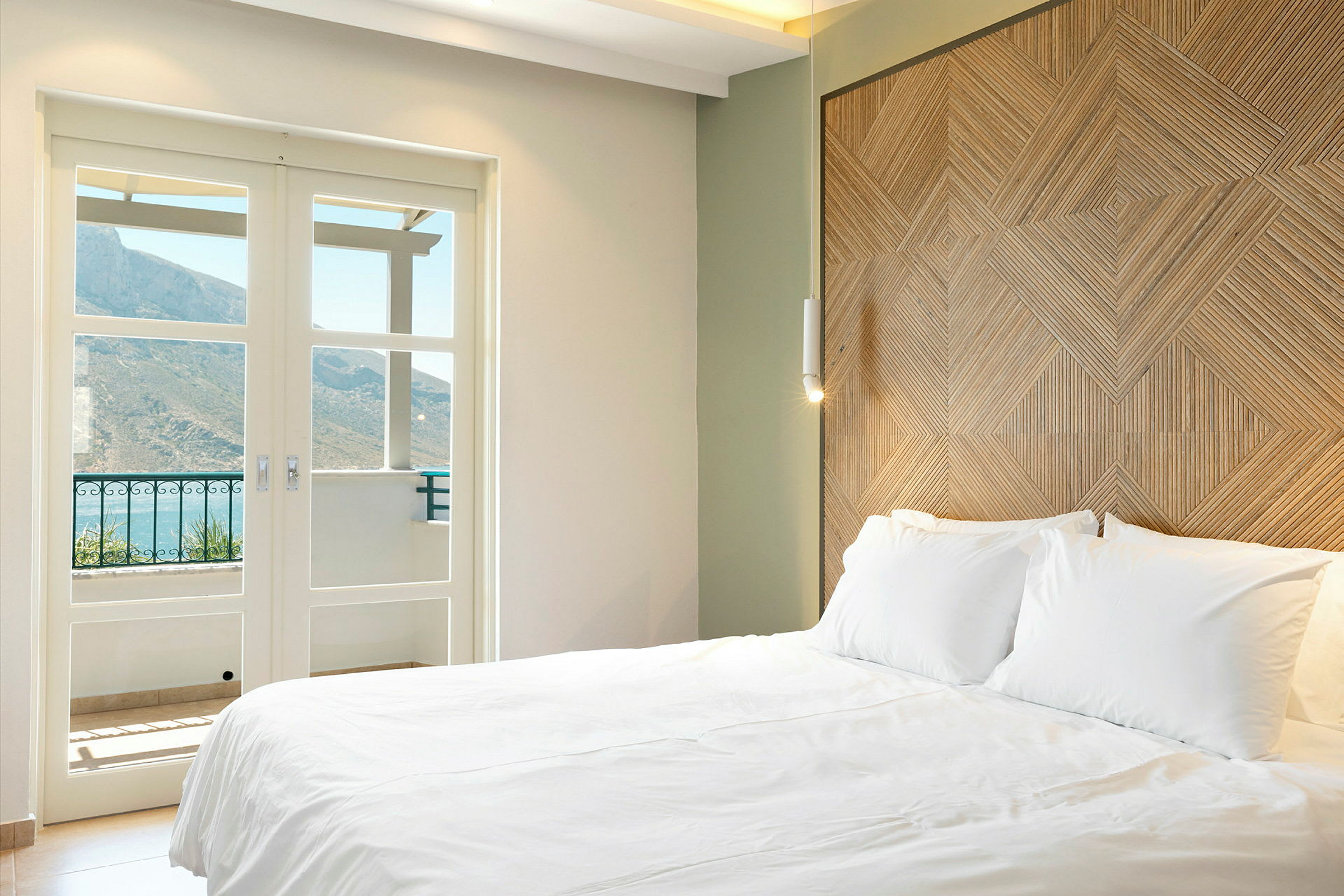 Ett hotellrum med en stor vit säng, ett dekorativt trämönstrat huvudgavel, och glasdörrar öppna mot en balkong med berg i bakgrunden. (AI-genererad text)