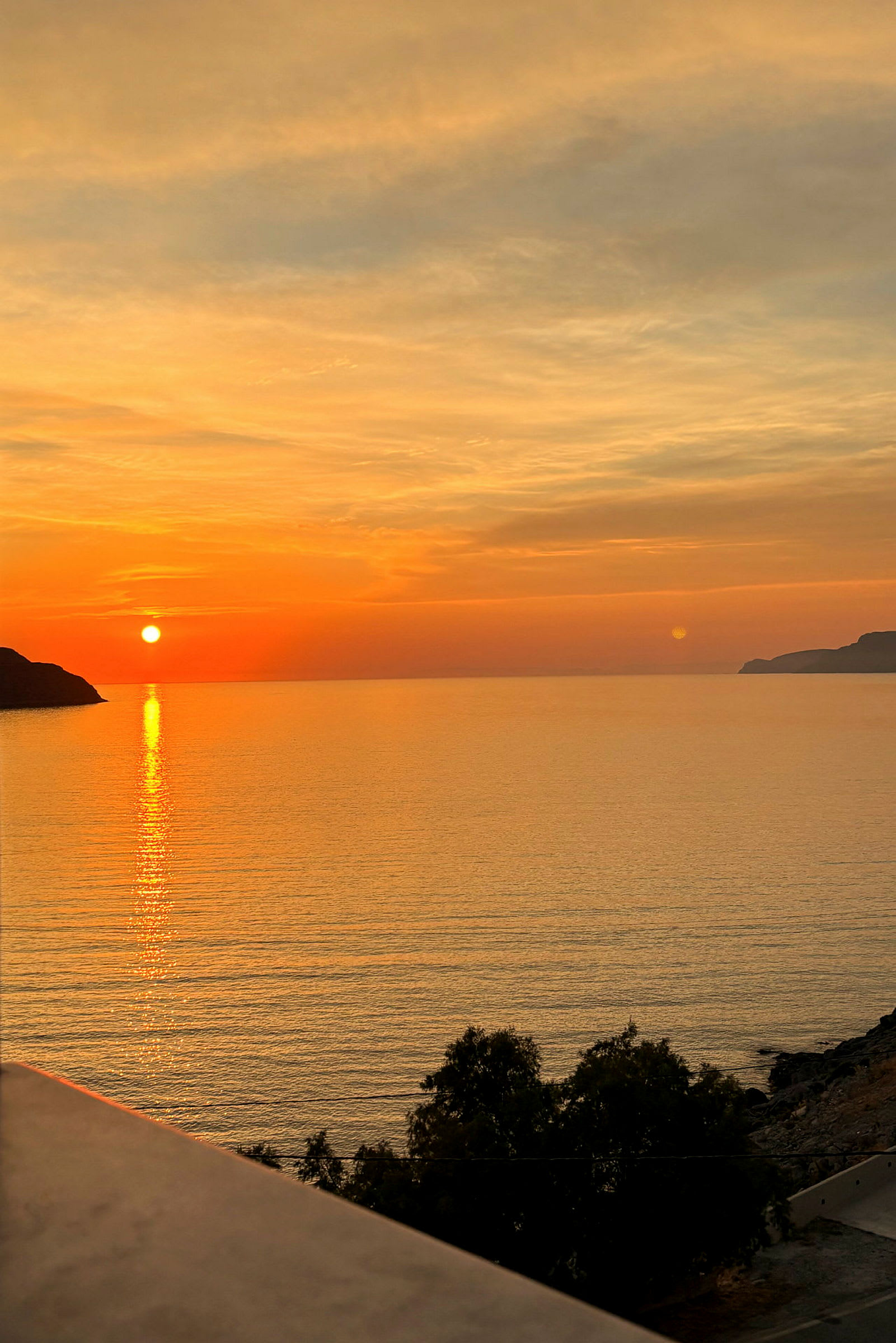 En solnedgång över havet, med orange och gula färger på himlen, några träd i förgrunden och en klippa till vänster. (AI-genererad text)