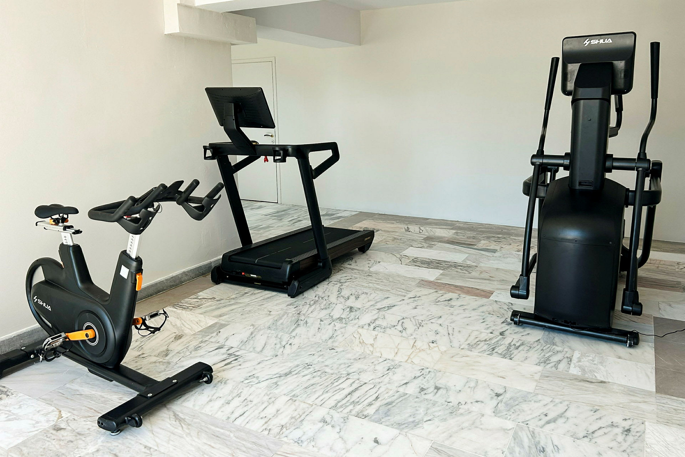 Bild på ett gym med en träningscykel, ett löpband och en crosstrainer placerade på ett golv med marmorklinker. (AI-genererad text)
