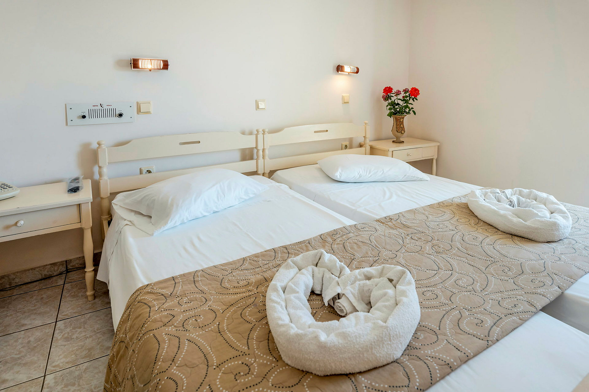 Ett hotellrum med två enkelsängar, prydnadsföremål och en vas med blommor på ett nattduksbord. (AI-genererad text)