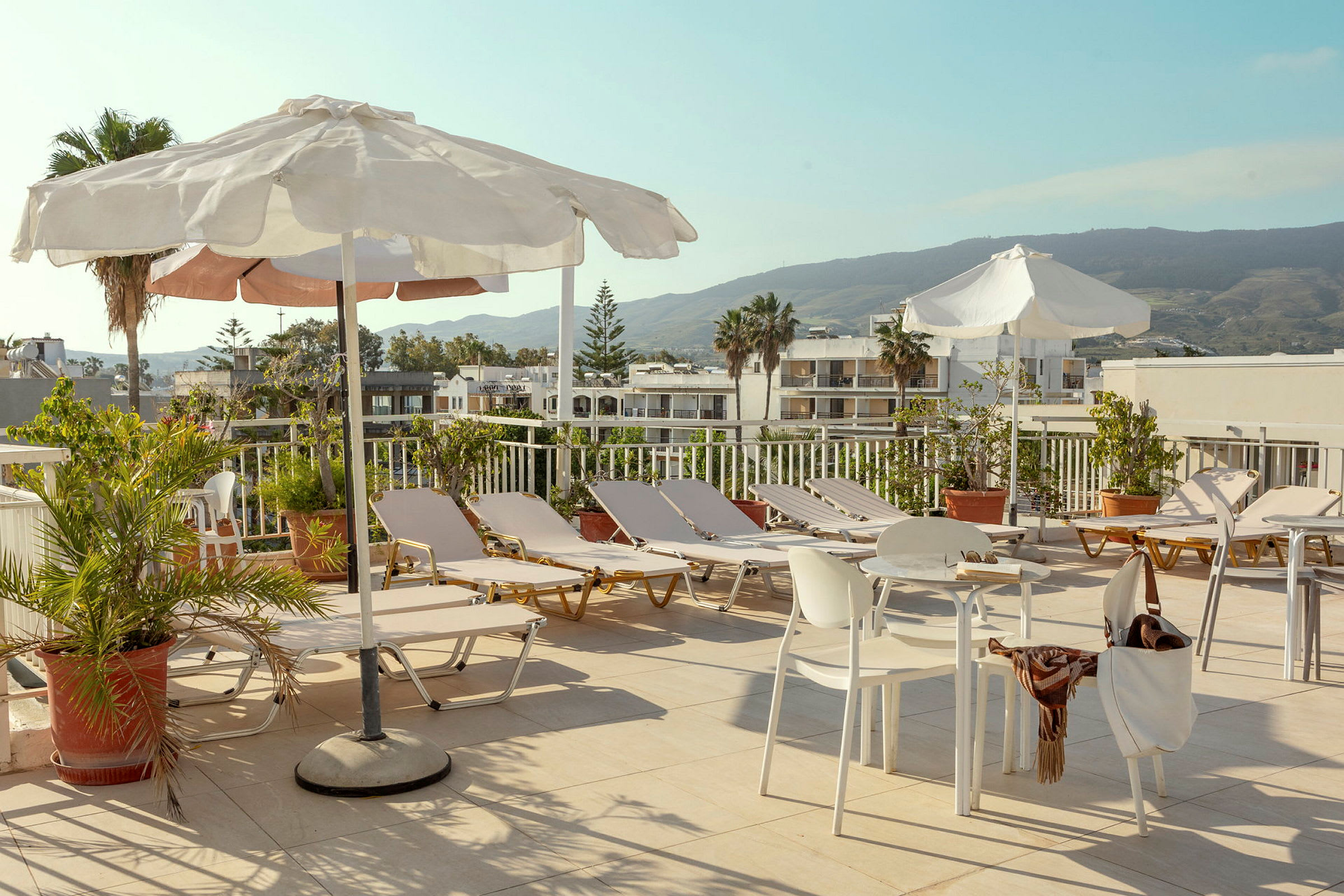 En solterrass med solstolar, parasoller och bord samt stolar omgivna av grönska och med utsikt över byggnader och berg. (AI-genererad text)