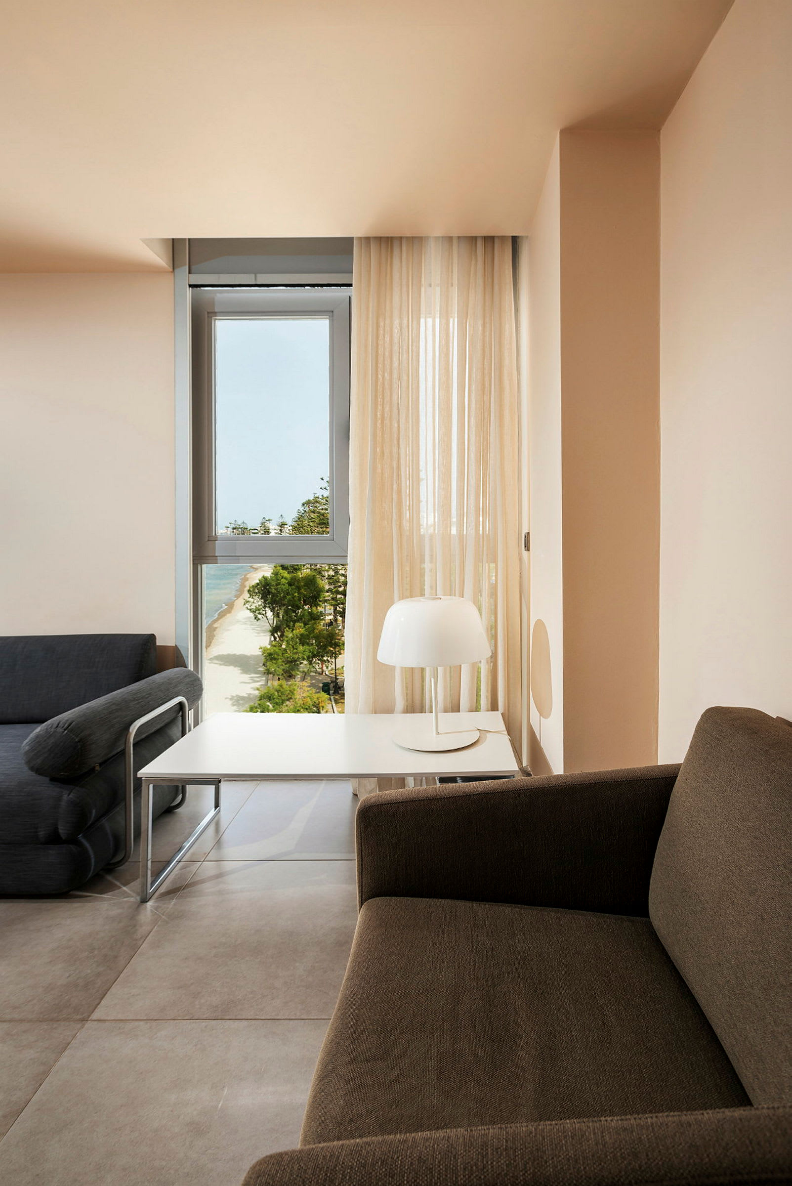 Ett rum med en soffa, ett bord och en lampa, med utsikt över en strand genom ett fönster. (AI-genererad text)