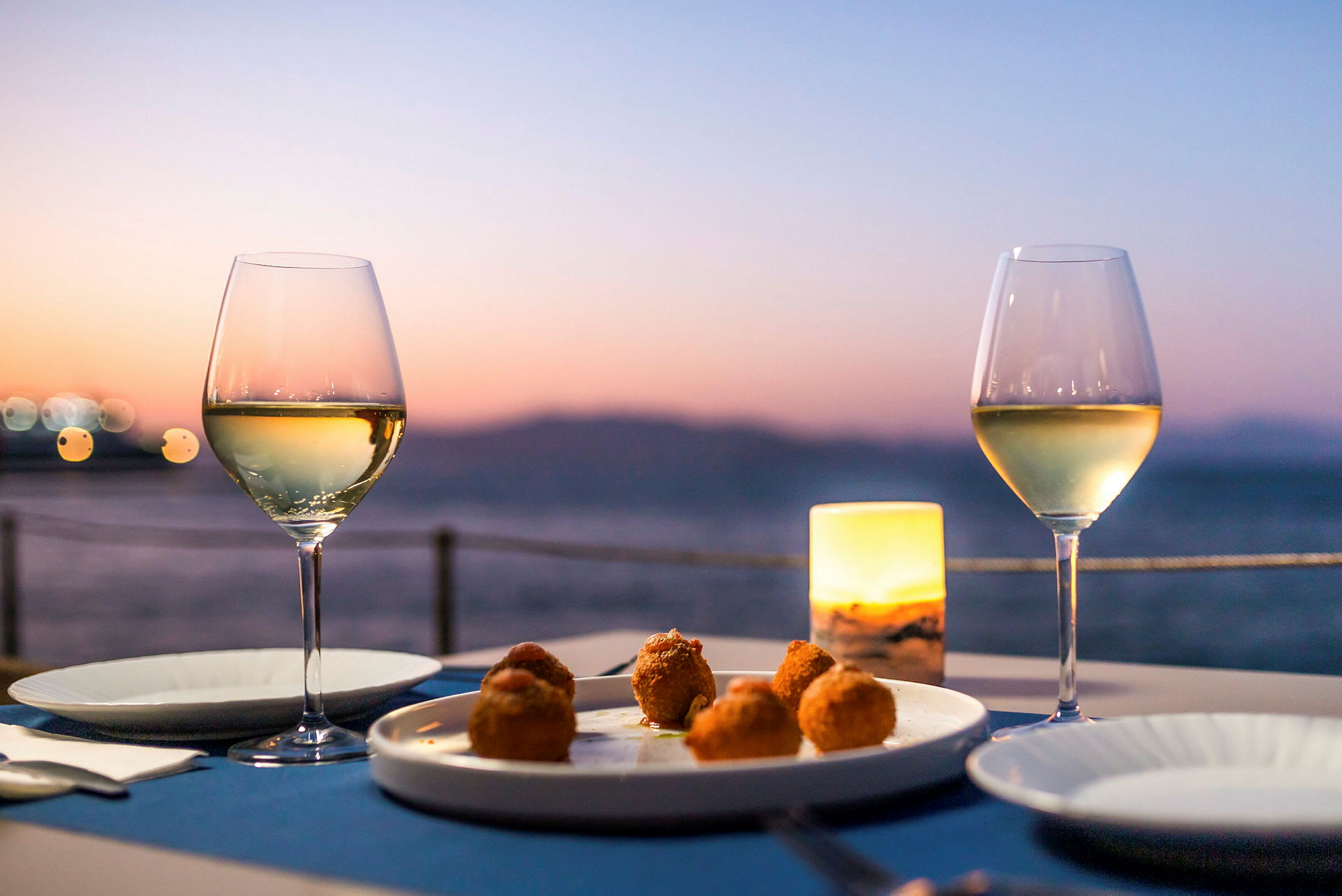 Två glas med vitt vin på ett dukat bord med en tallrik med mat framför en solnedgång vid havet. (AI-genererad text)