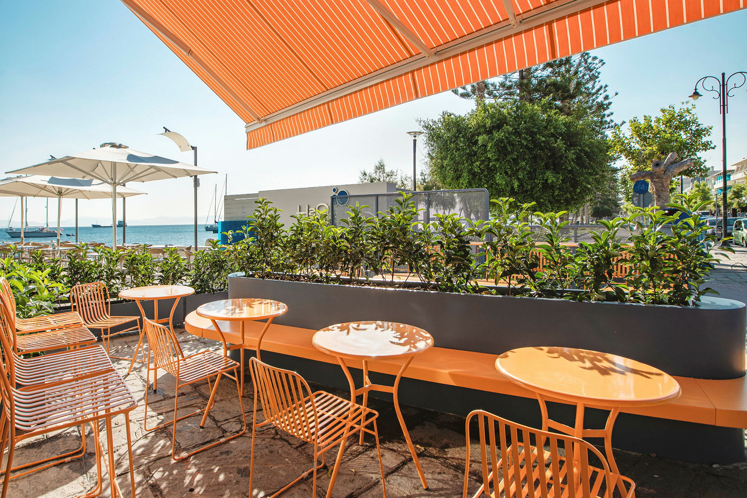 En uteservering med orange bord och stolar samt vita parasoller mot bakgrunden av havet. (AI-genererad text)