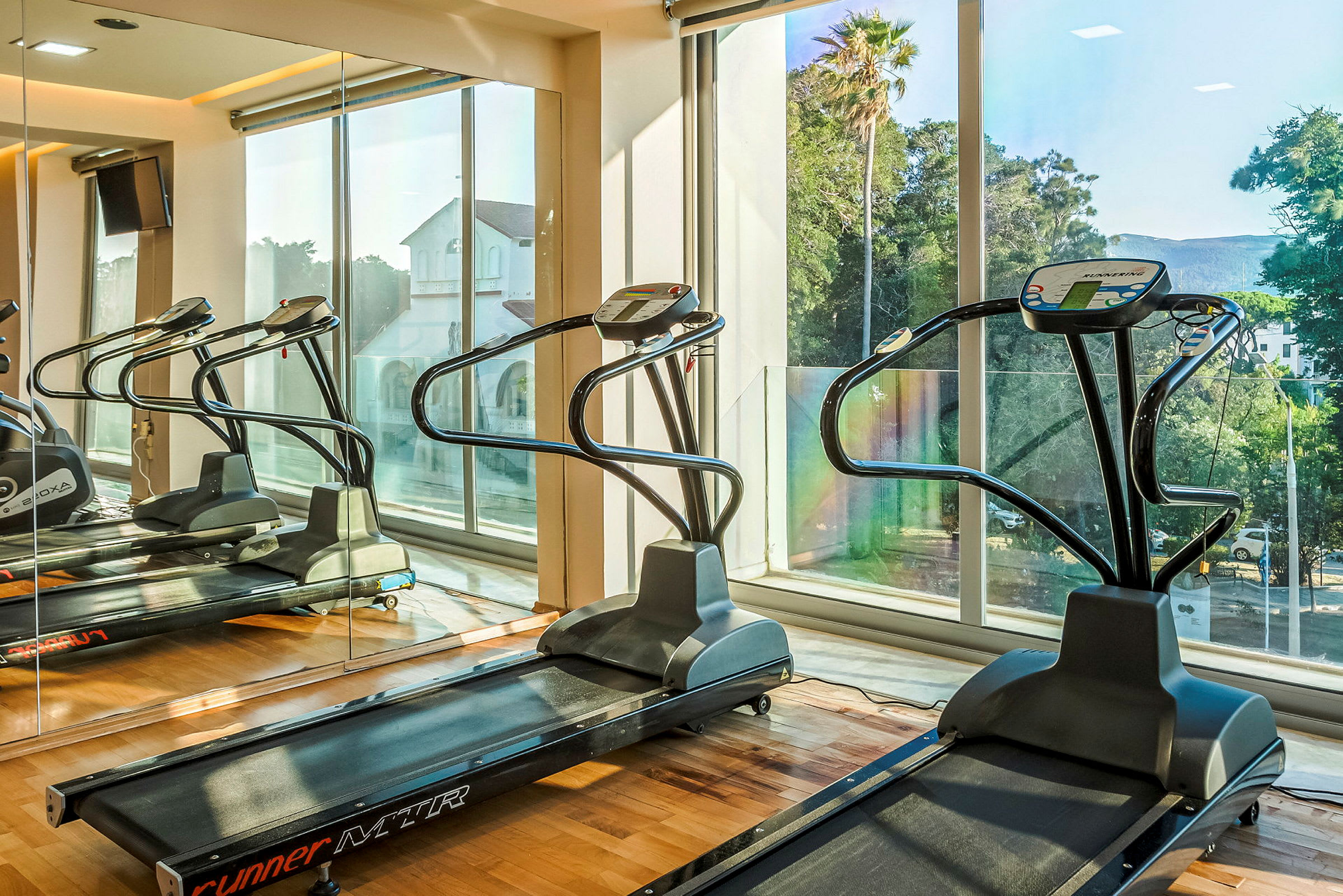 Ett gym med löpband framför stora fönster med utsikt över träd och byggnader. (AI-genererad text)