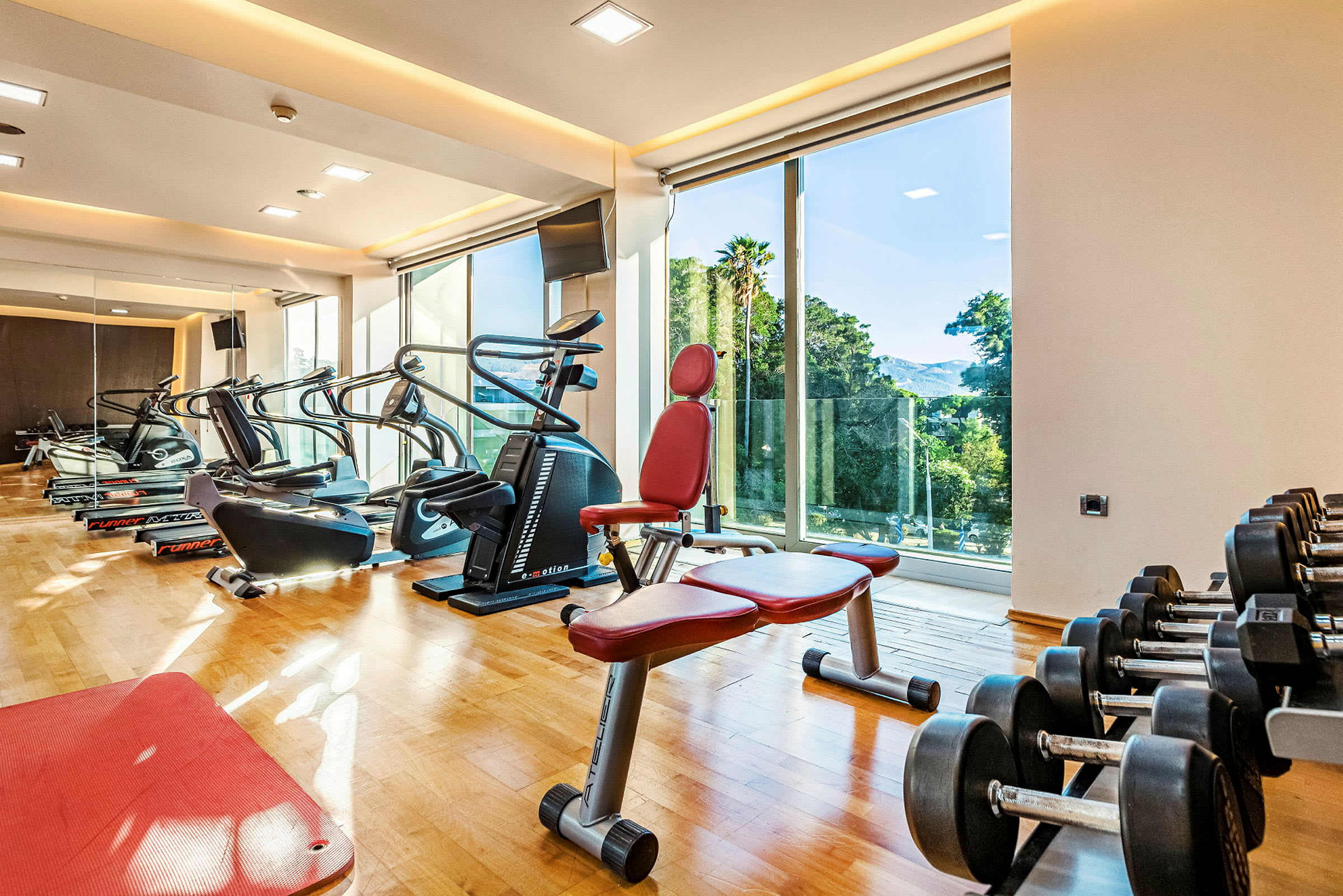 Ett gym med träningsmaskiner, hantlar och bänkar, beläget i ett ljust rum med stora fönster som visar utsikt över naturen. (AI-genererad text)