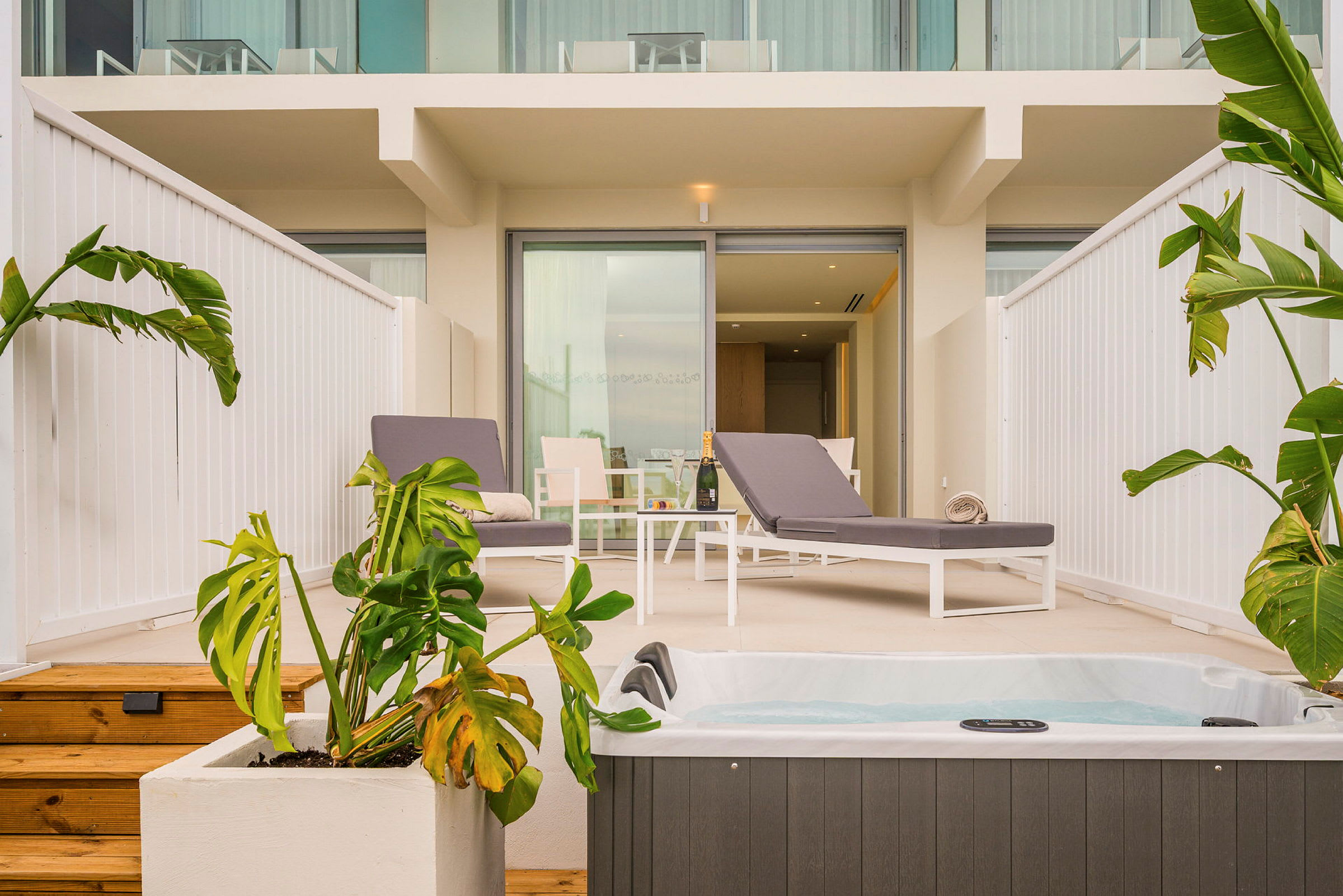 En uteterrass med två solstolar, ett bord och en jacuzzi i förgrunden. Det finns gröna växter runtom. (AI-genererad text)