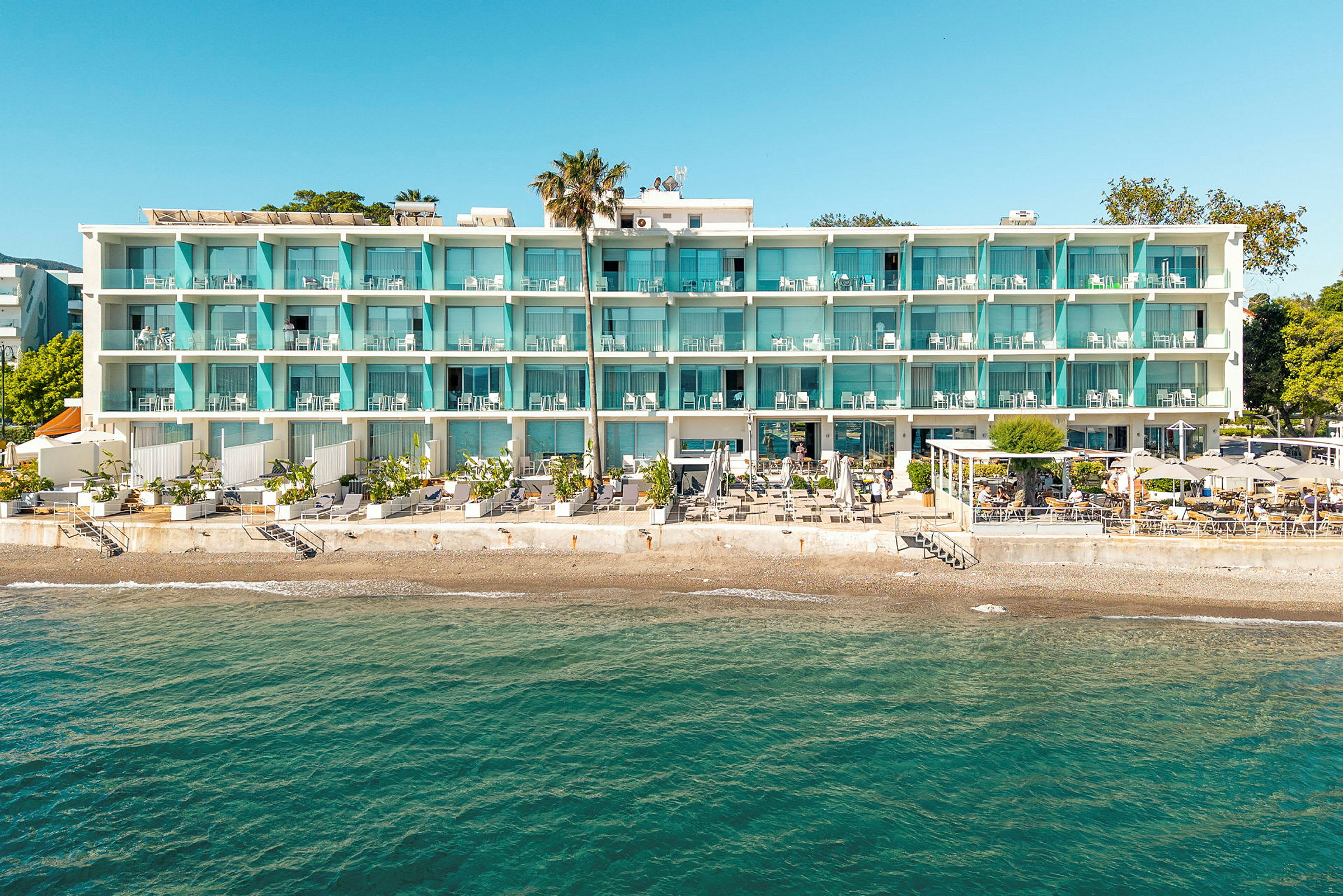 Ett hotell vid stranden med en vit fasad och blå balkonger. Det finns en terrass med parasoller och stolar nedanför, och havet syns i förgrunden. (AI-genererad text)