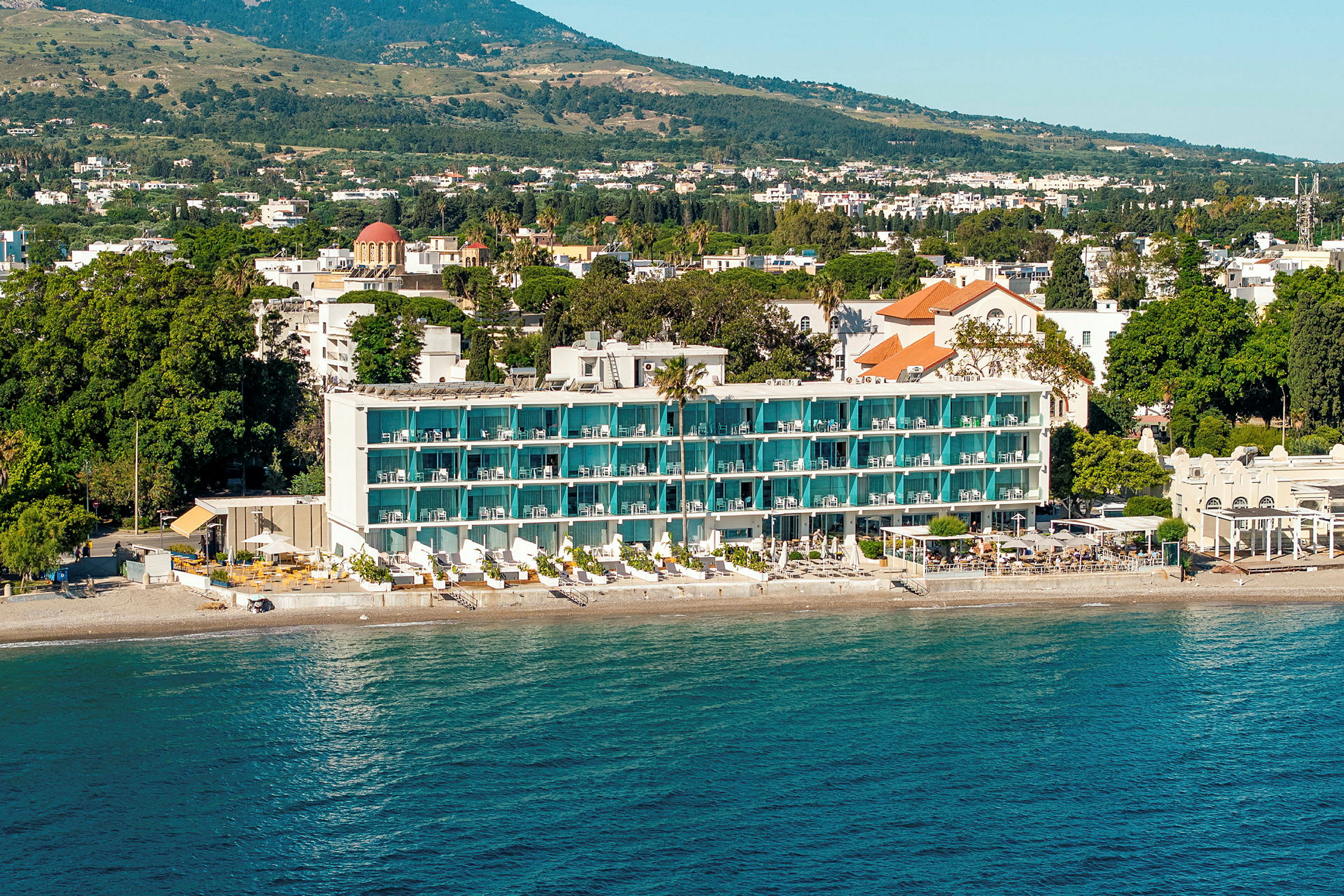 En hotellbyggnad med balkonger och en strand framför, omgivna av gröna träd och ett landskap i bakgrunden. (AI-genererad text)