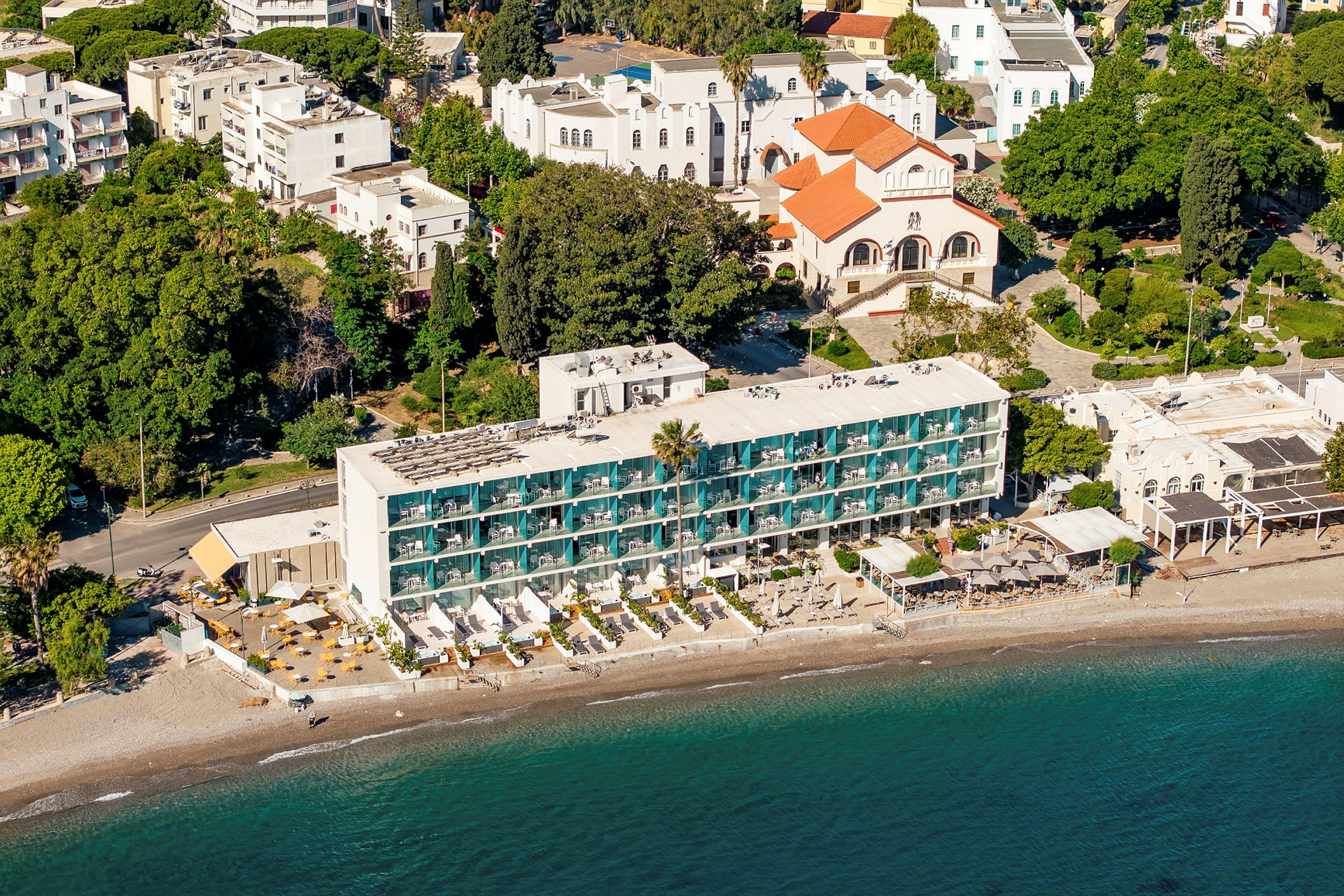 Flygfoto av en hotellbyggnad vid en strand, omgiven av grönska och andra byggnader. (AI-genererad text)