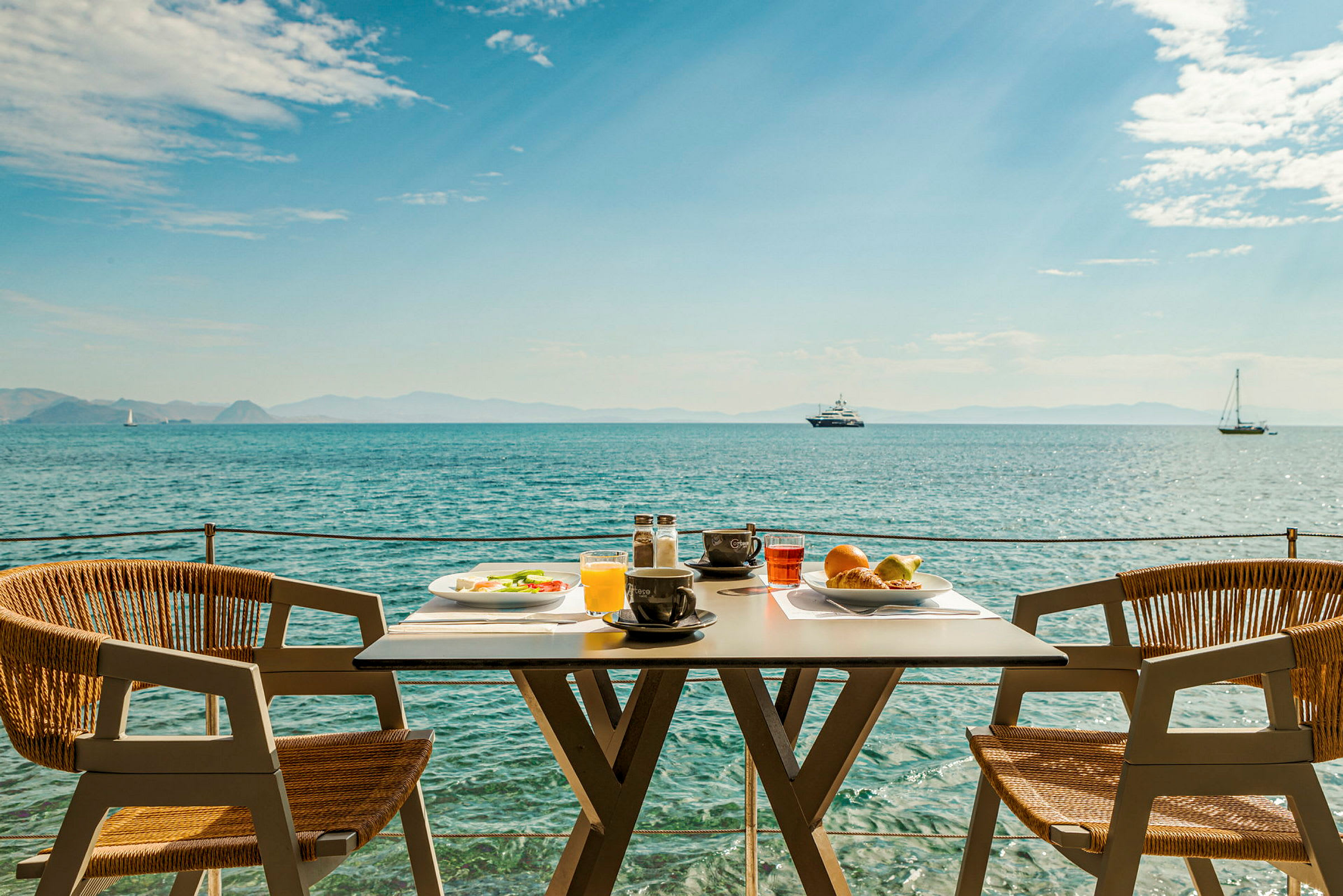 Ett dukat bord med frukost står vid havet med båtar i bakgrunden. (AI-genererad text)
