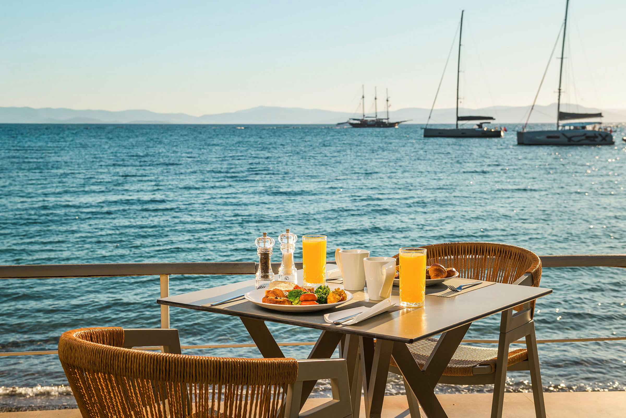 Ett bord med frukost och juice står vid havet, med segelbåtar i bakgrunden. (AI-genererad text)