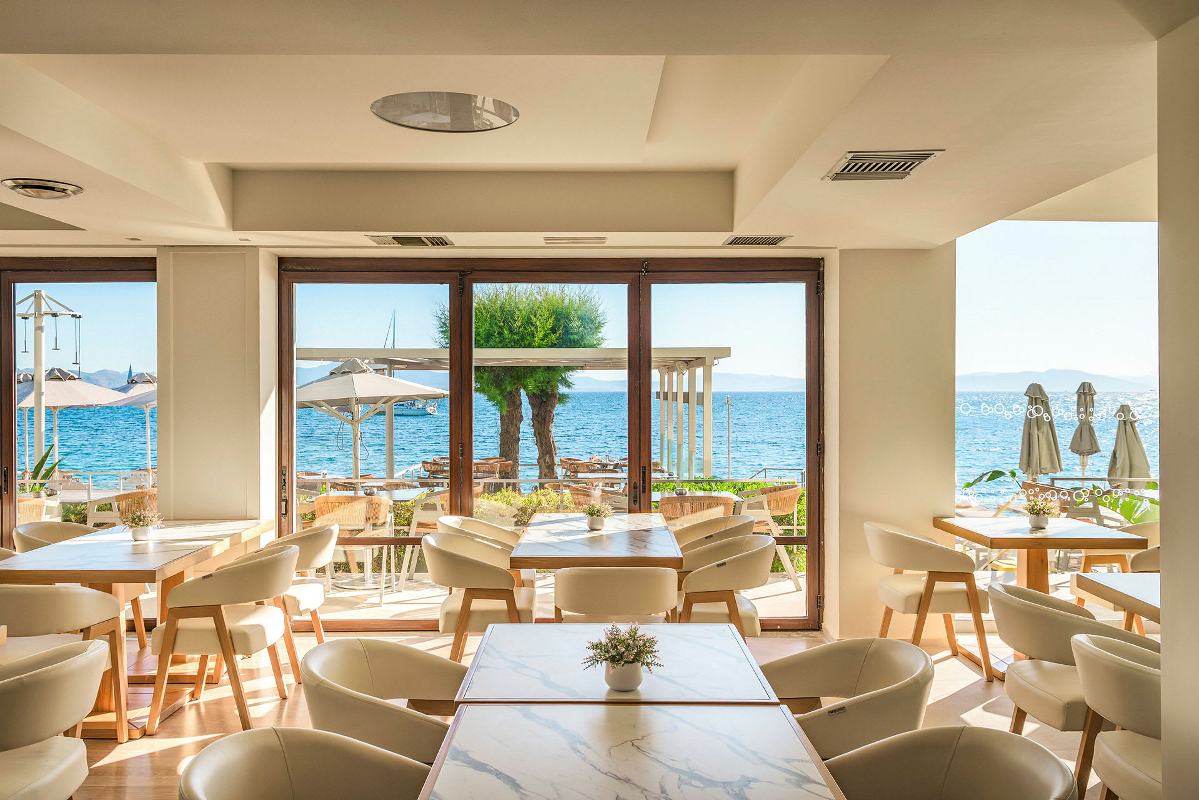 Ett ljus och modern restaurang med utsikt över havet genom stora fönster. Bord med stolar står inne, och utanför syns parasoller samt vatten. (AI-genererad text)