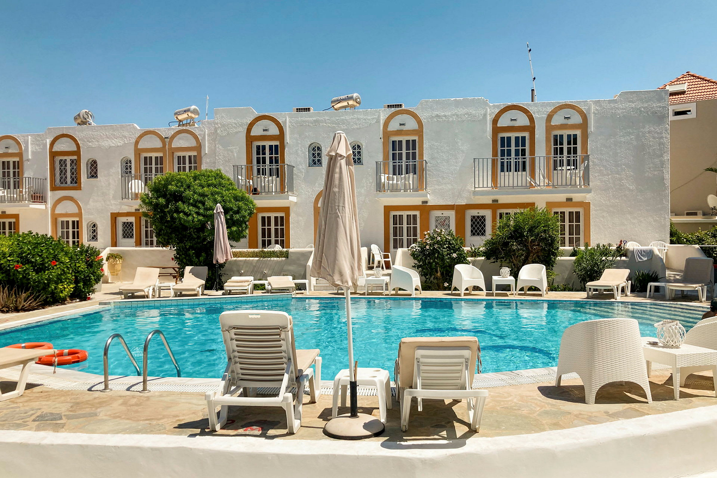 En hotellpool med solstolar och parasoll framför en byggnad med vita fasader och orange detaljer. (AI-genererad text)