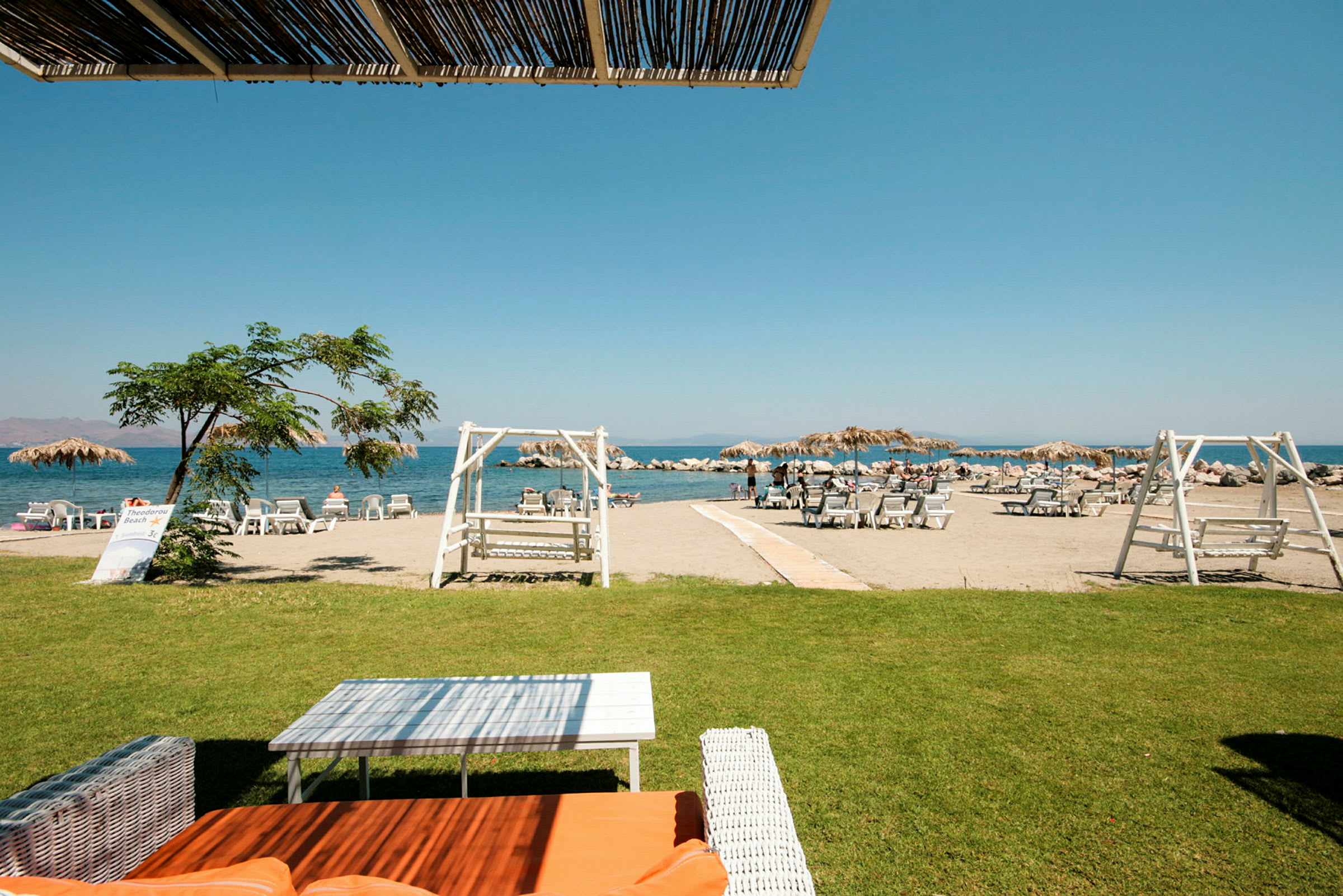 Strand med solstolar och parasoller nära havet, gräsmatta och trämöbler i förgrunden. (AI-genererad text)