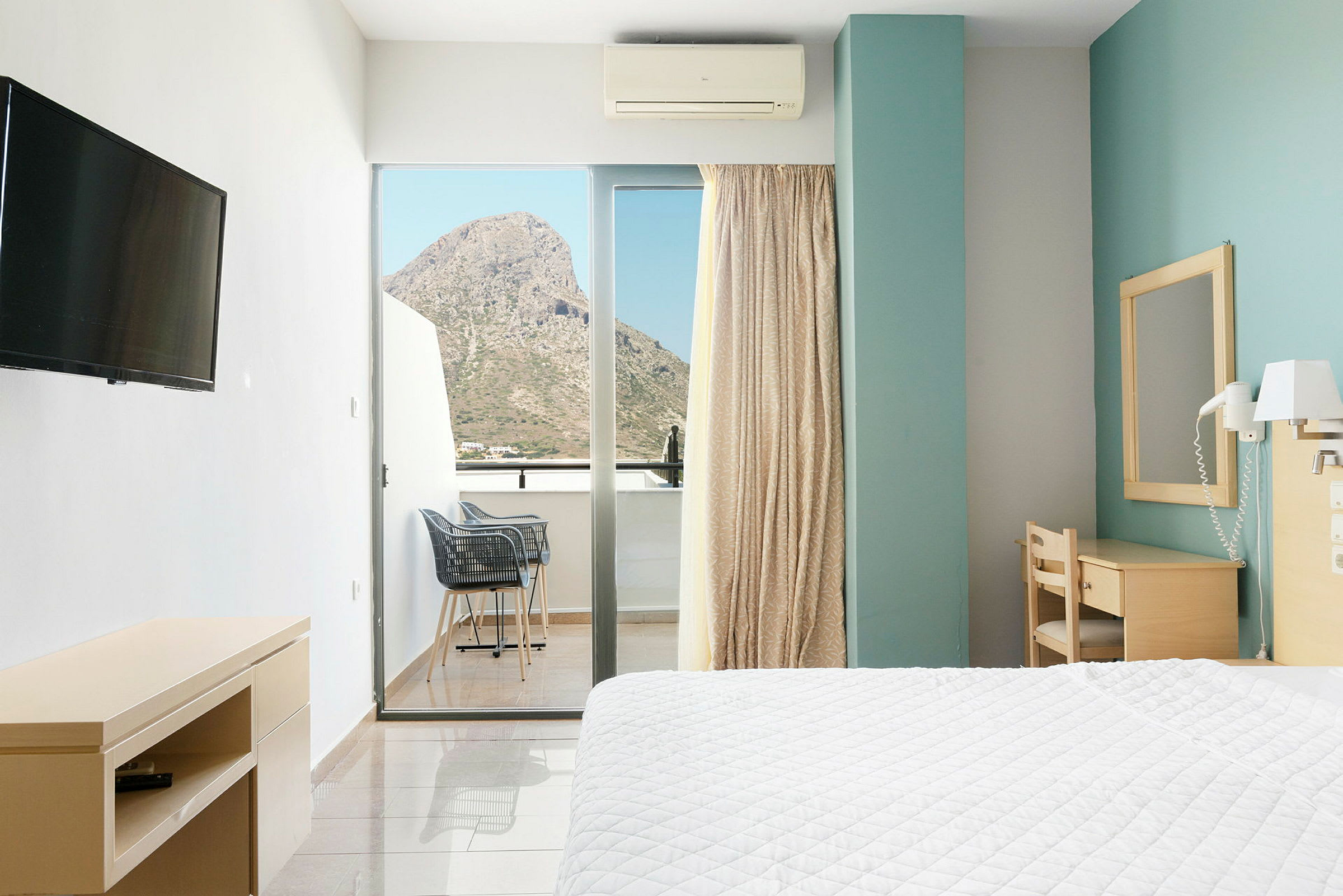 Hotellrum med en säng, skrivbord och balkong med utsikt över ett berg. (AI-genererad text)
