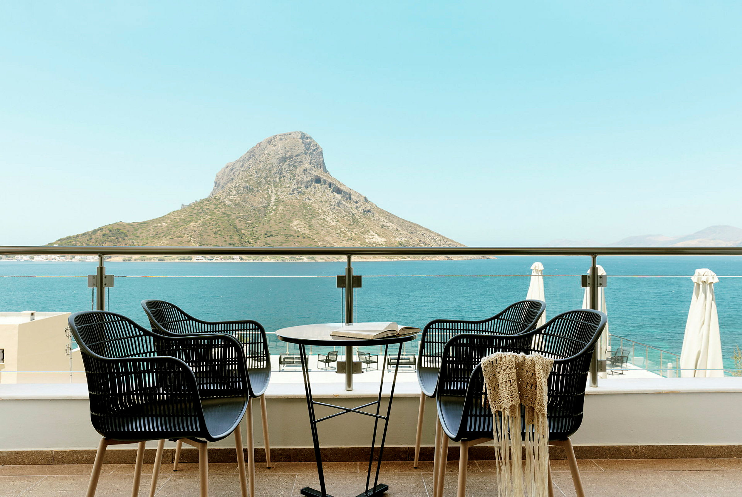 Fyra stolar och ett bord på en terrass med utsikt över havet och en bergstopp. (AI-genererad text)