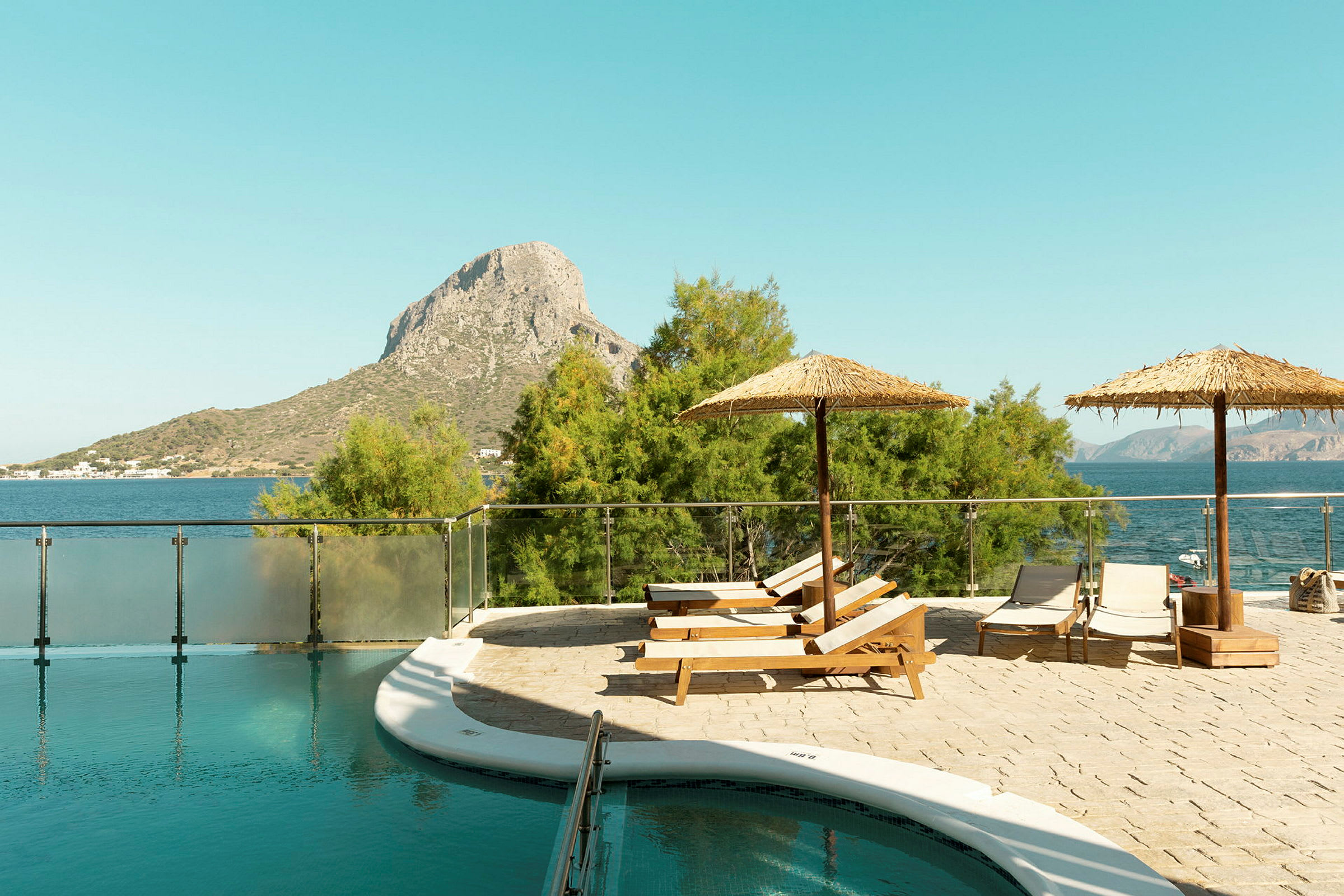 En pool med solstolar och parasoller nära havet och en bergskulle i bakgrunden. (AI-genererad text)