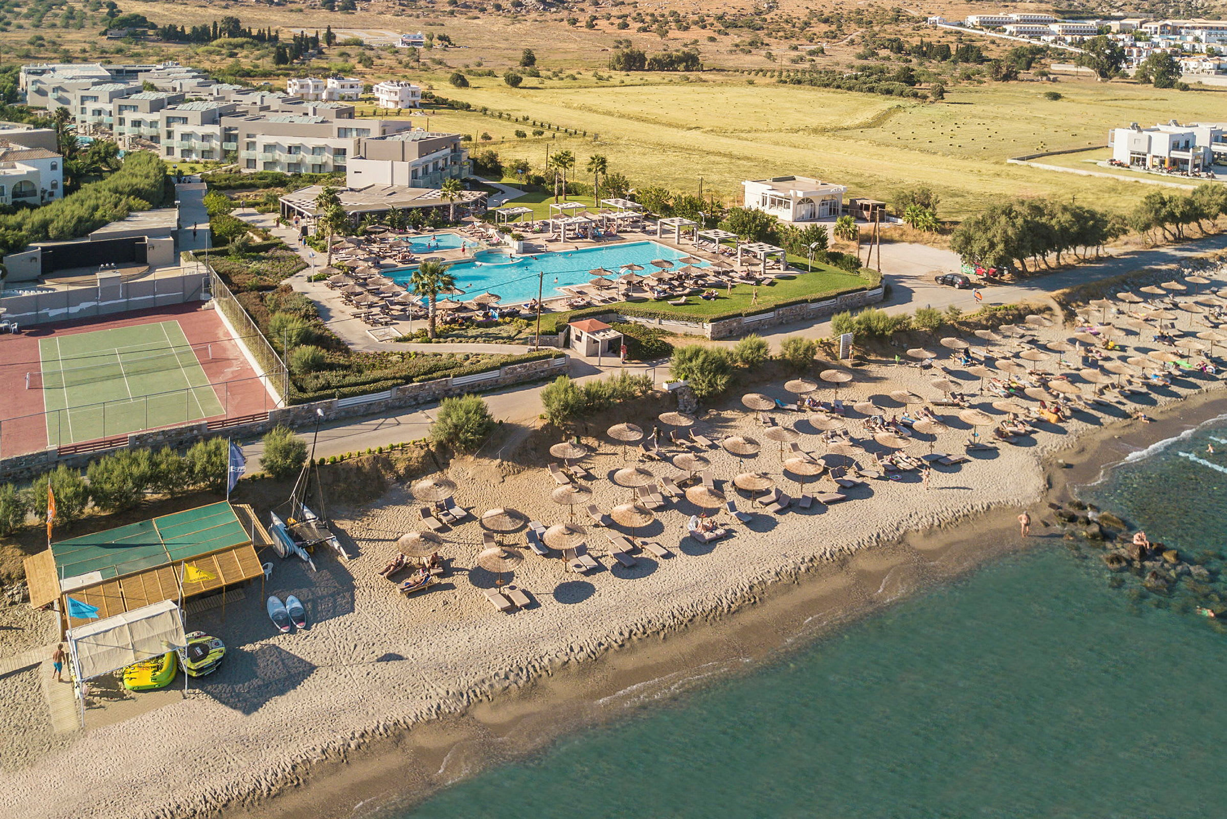 Flygbild av en strand med solstolar och parasoller, en pool samt byggnader i bakgrunden. (AI-genererad text)