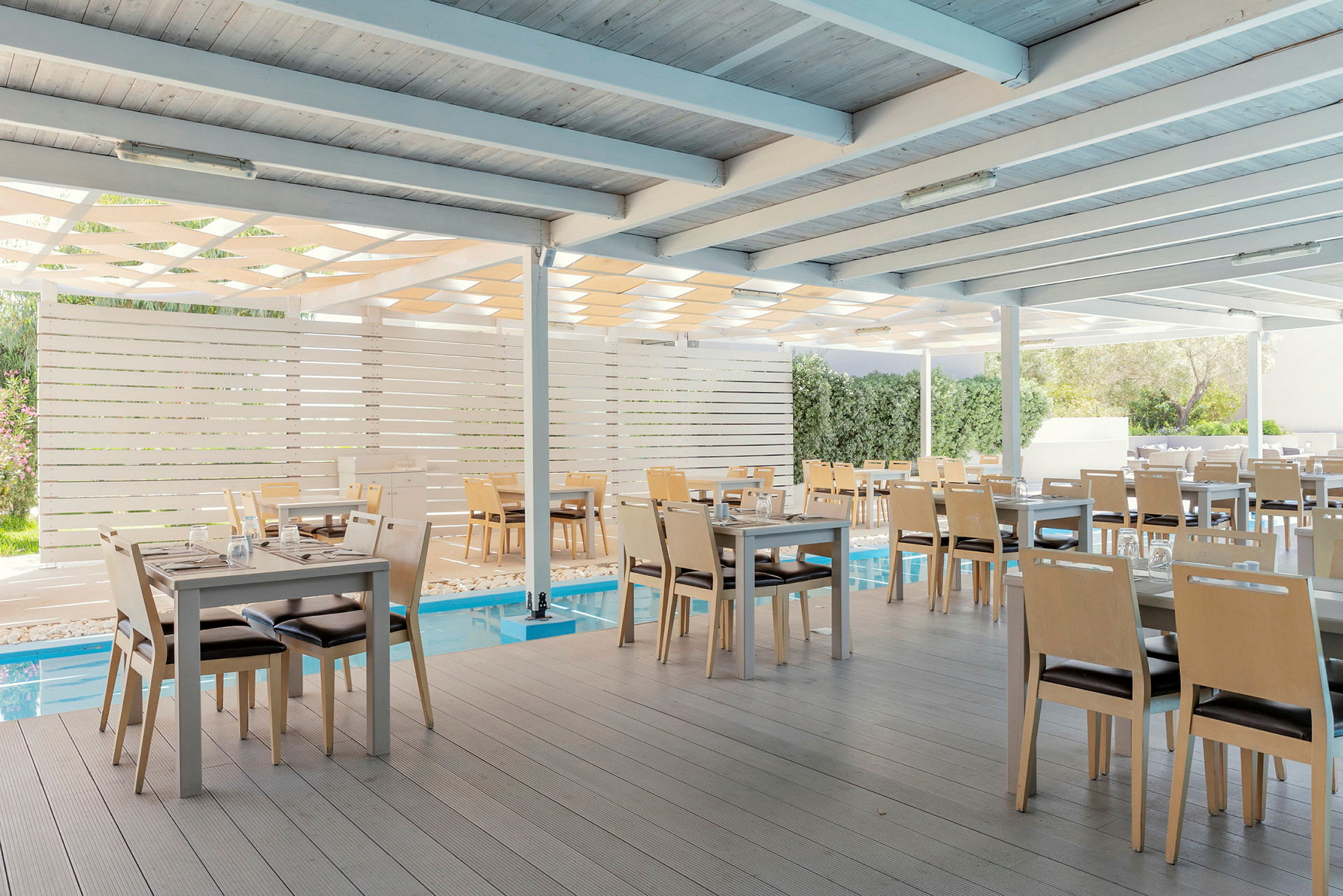 En täckt matplats med bord och stolar vid en pool, omgiven av ljusa träpaneler och gröna buskar. (AI-genererad text)