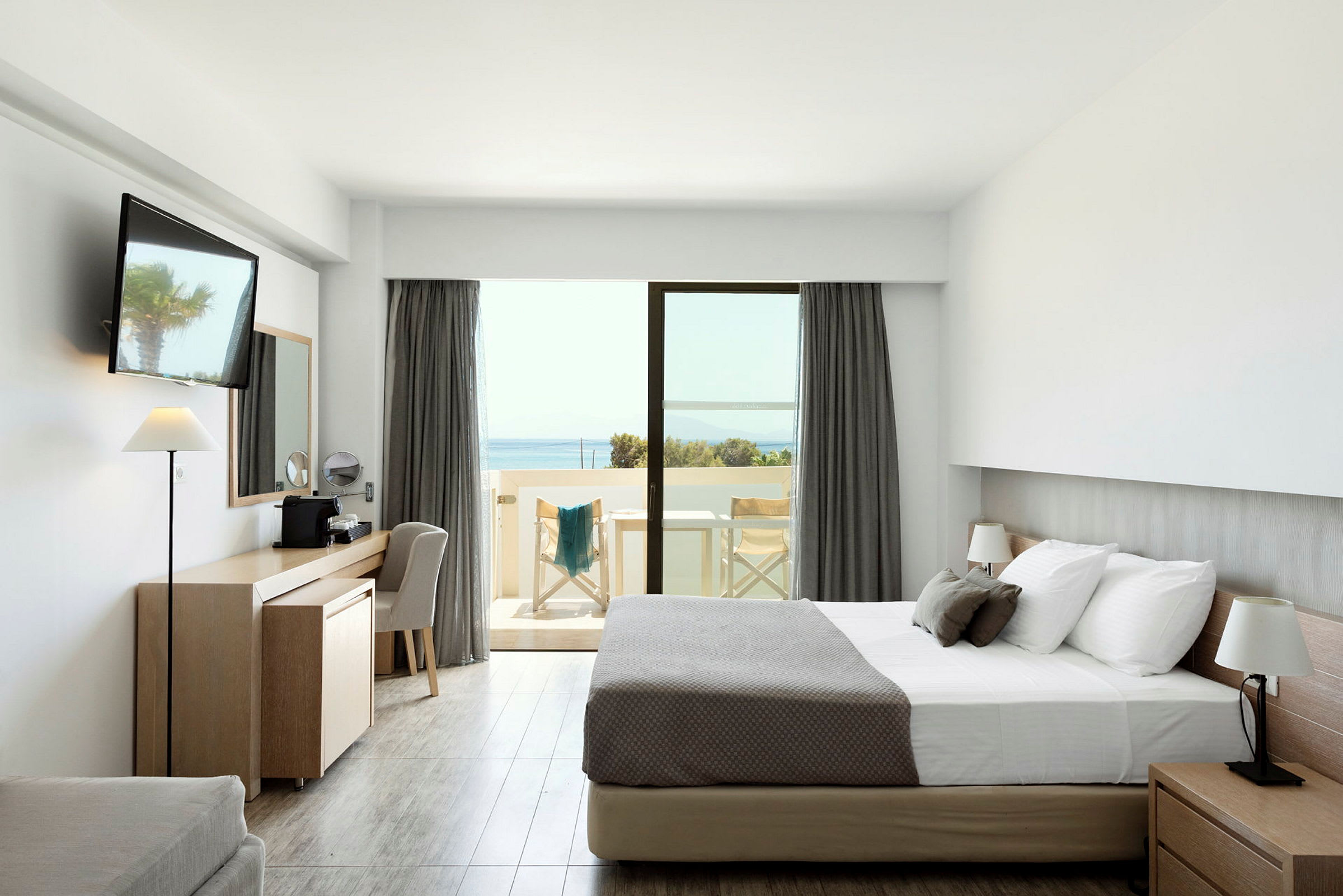 En hotellrum med en dubbelsäng, skrivbord, och en balkong med utsikt över havet. (AI-genererad text)