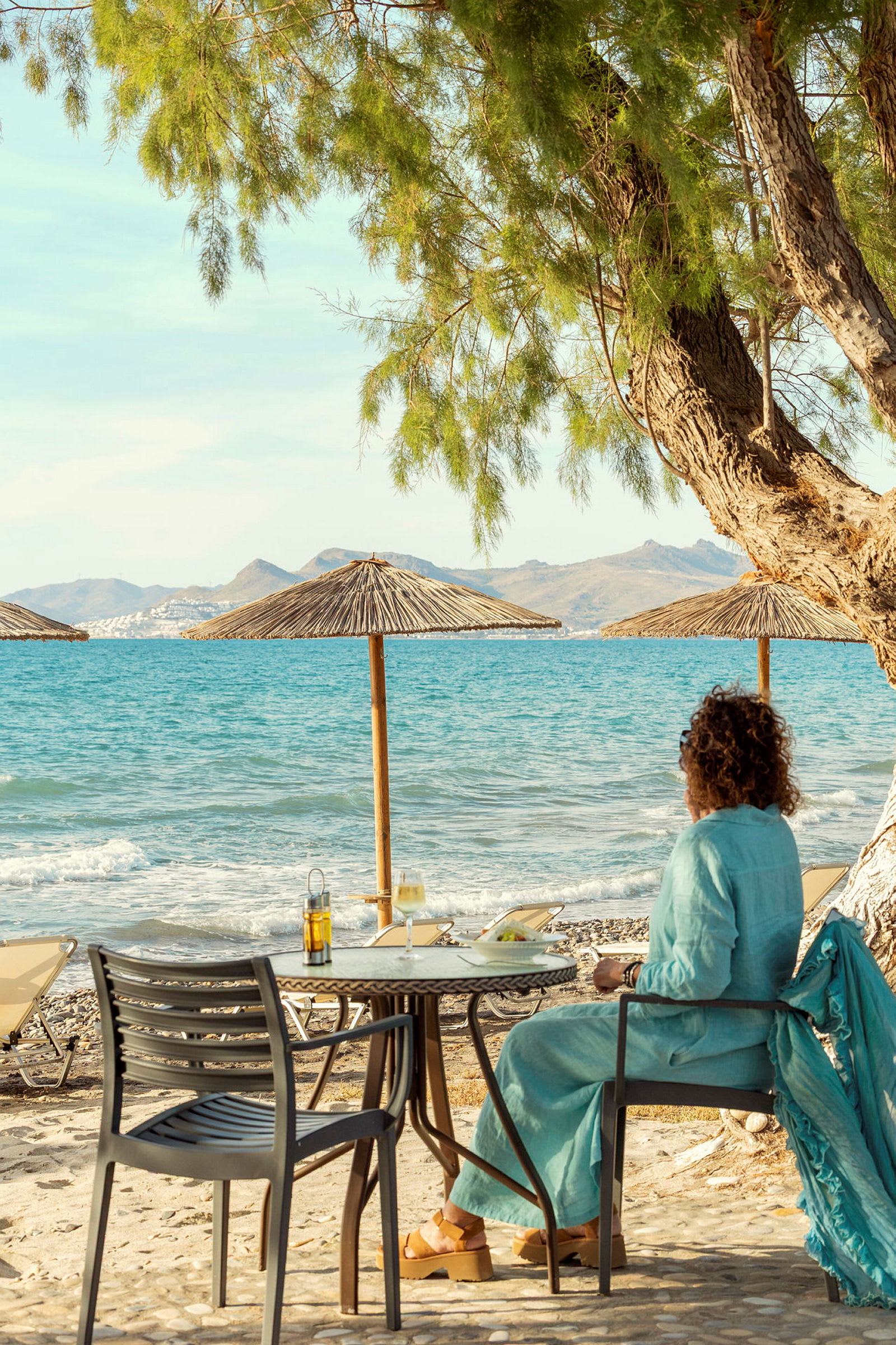 Person sitter vid ett bord på en stenstrand med utsikt över havet och berg i bakgrunden. (AI-genererad text)