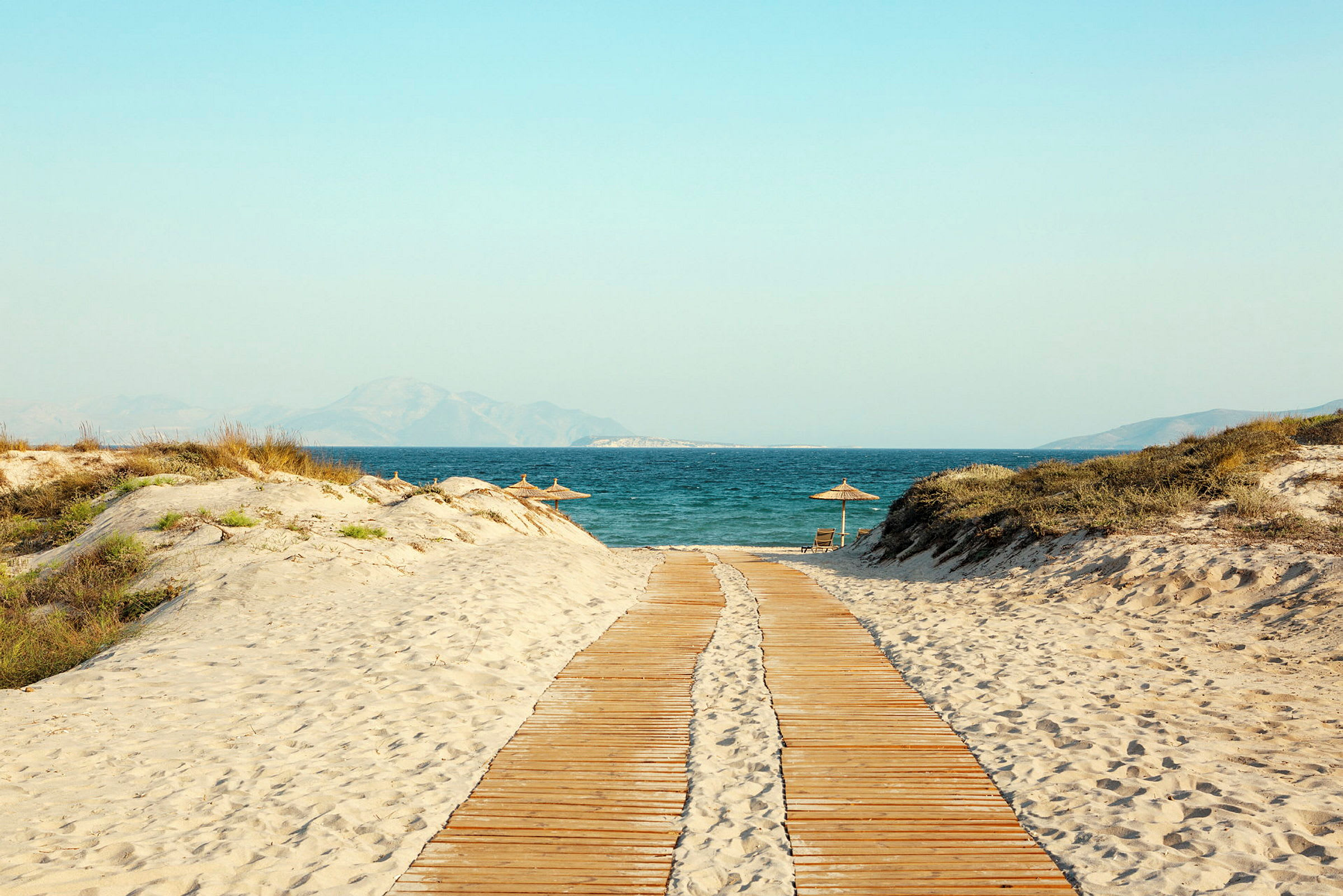 En träbrygga leder över en sandstrand till havet. (AI-genererad text)