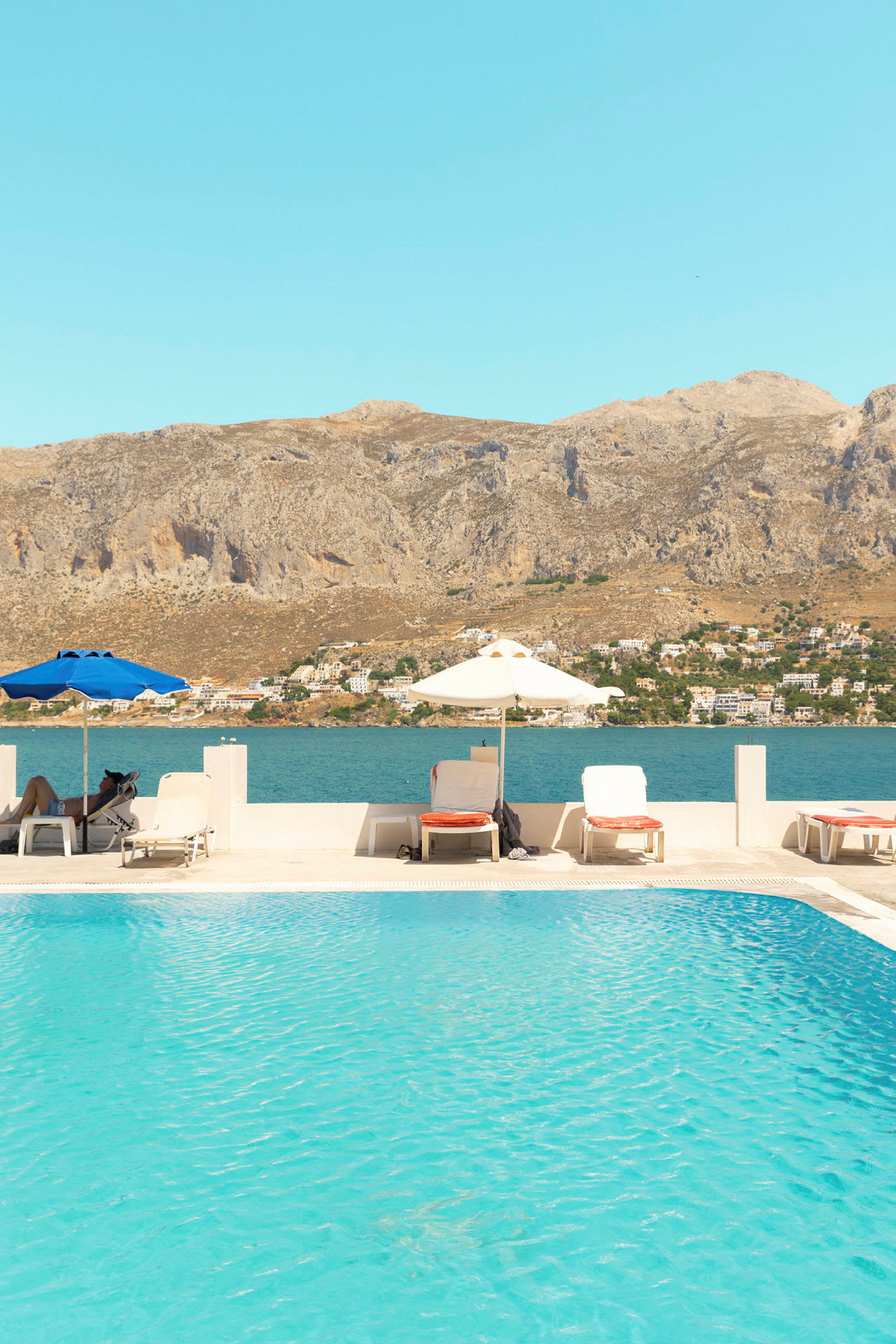 En swimmingpool med solsängar och parasoller, med utsikt över havet och berg i bakgrunden. (AI-genererad text)