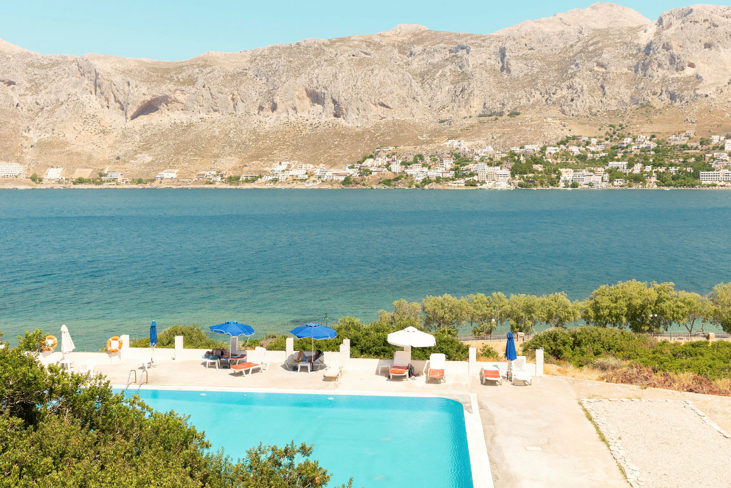 Utsikt över en pool med solstolar och parasoller, med havet och berg i bakgrunden. (AI-genererad text)