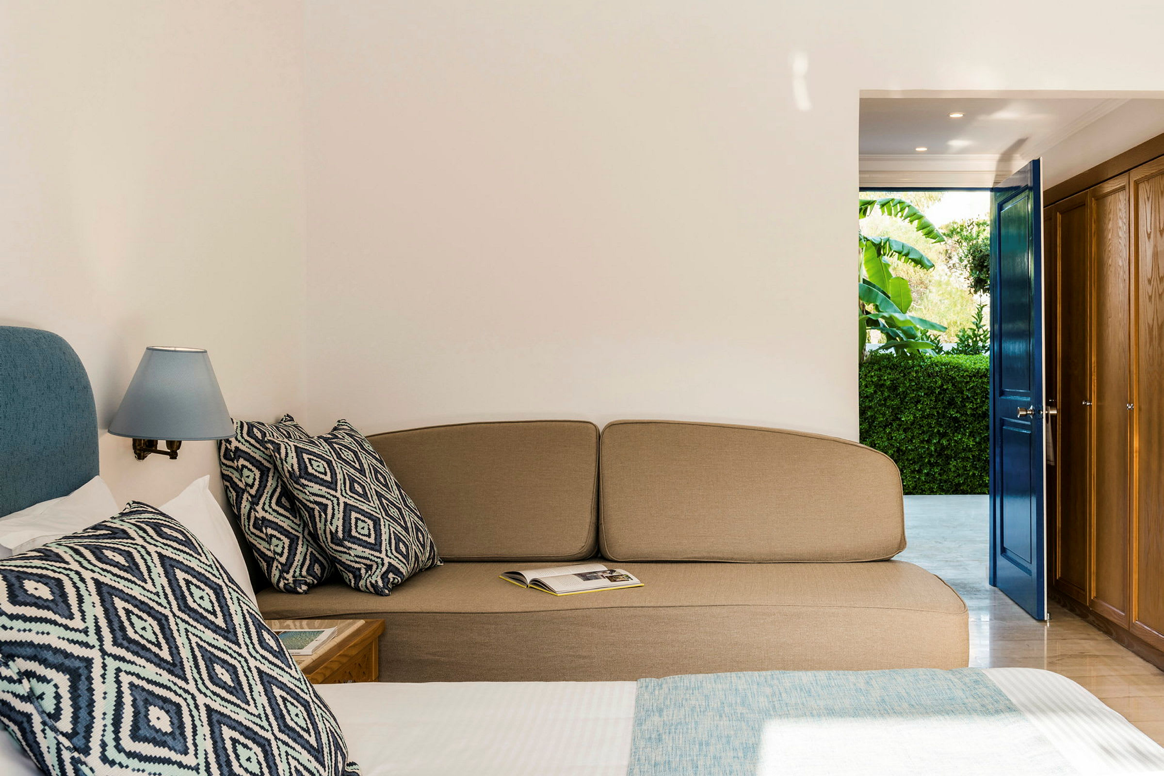 Hotellrum med en soffa, en säng med dekorativa kuddar och en öppen dörr mot en grönskande trädgård. (AI-genererad text)