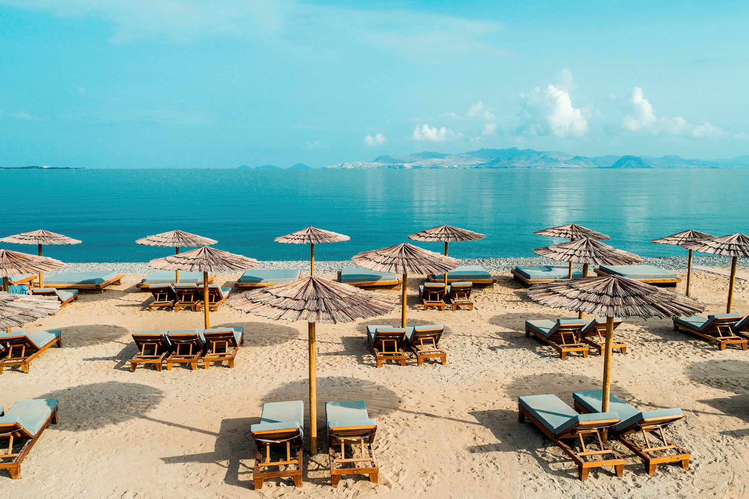Solstolar och parasoller på en sandig strand med klart vatten och berg i bakgrunden. (AI-genererad text)