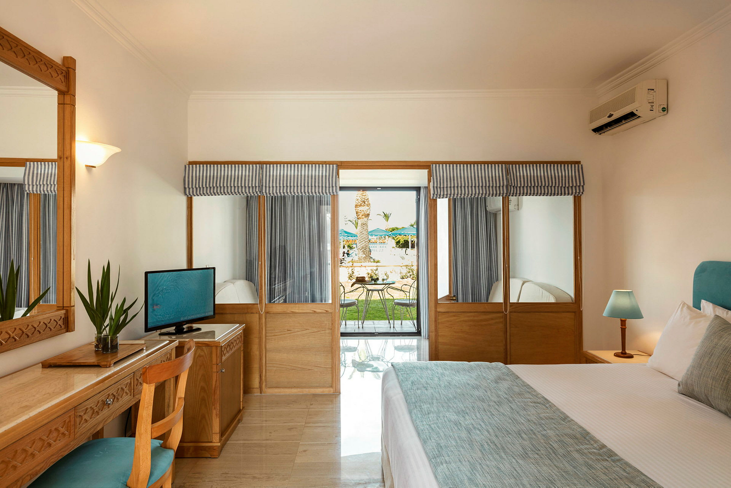 Ett hotellrum med en säng, ett skrivbord med en stol, en TV och ett fönster med utsikt mot en terrass och en trädgård utanför. (AI-genererad text)