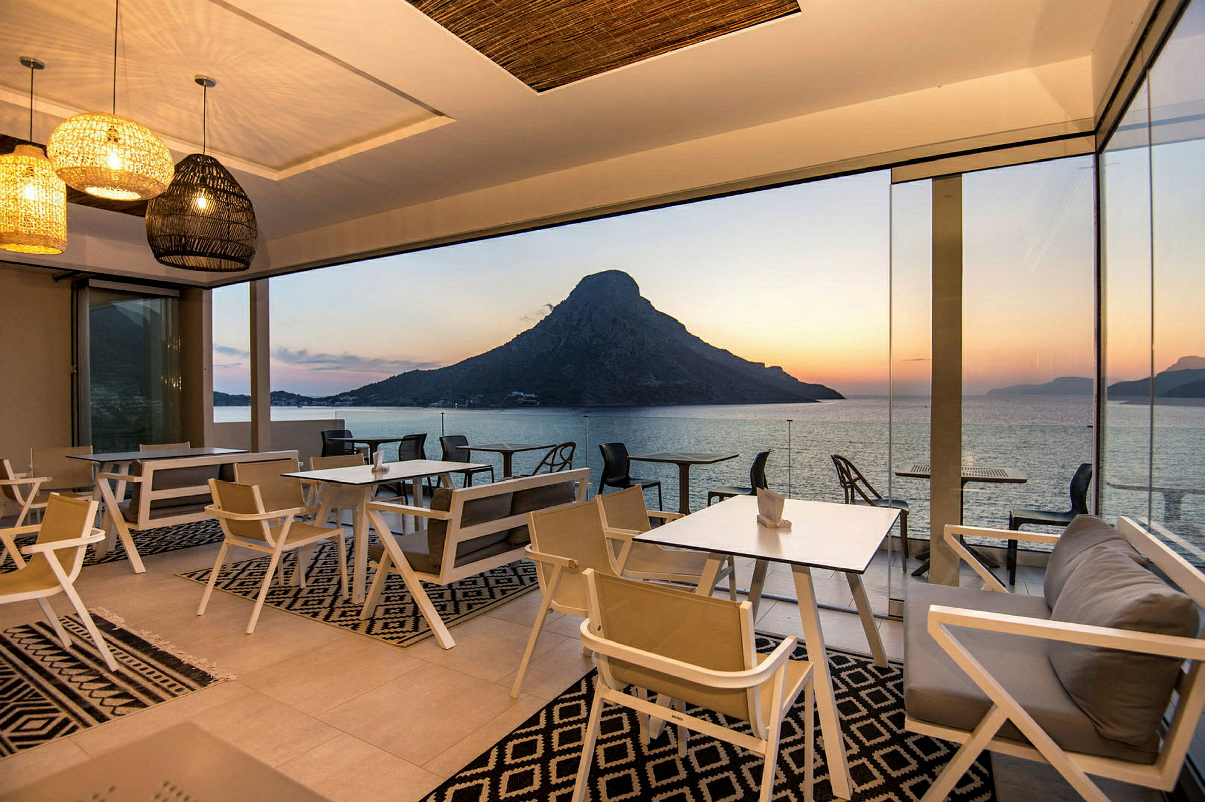 Restaurang med glasväggar och utsikt över havet och berg i solnedgång. (AI-genererad text)