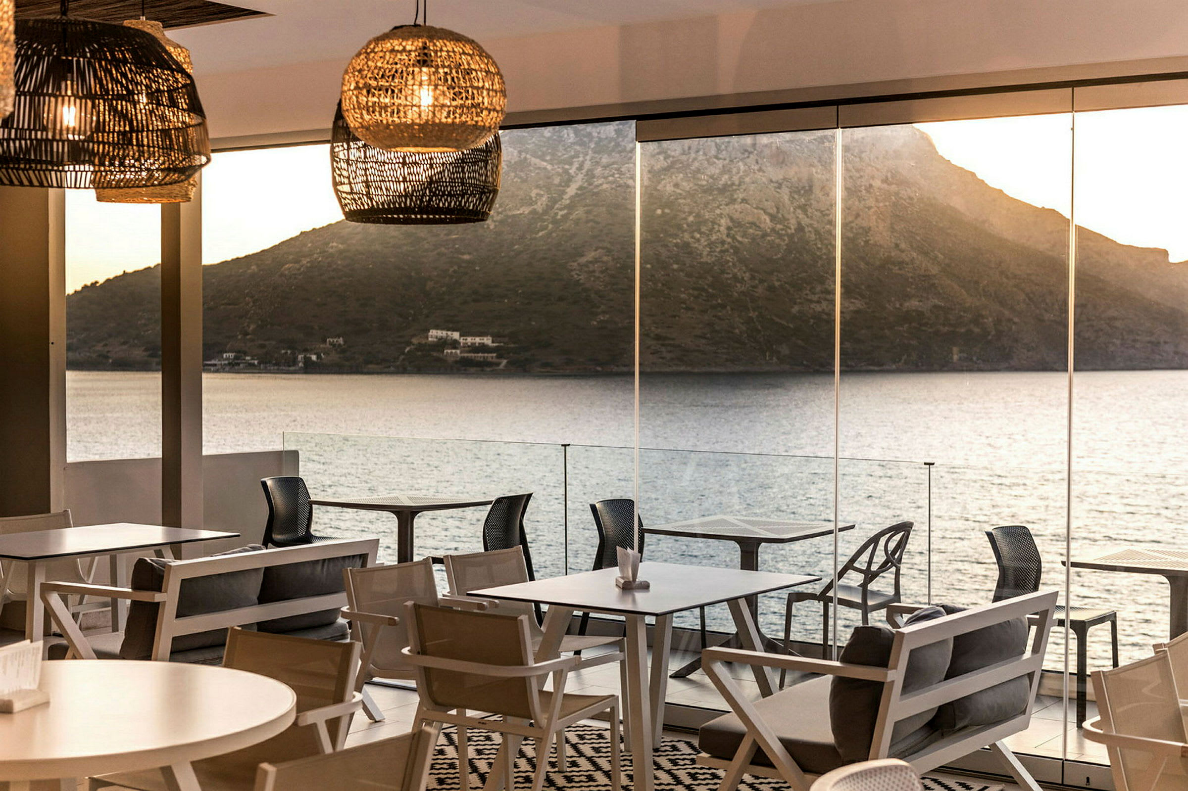 Utsikt över hav och berg bakgrunden från en restaurang med stora glasfönster och moderna möbler. (AI-genererad text)