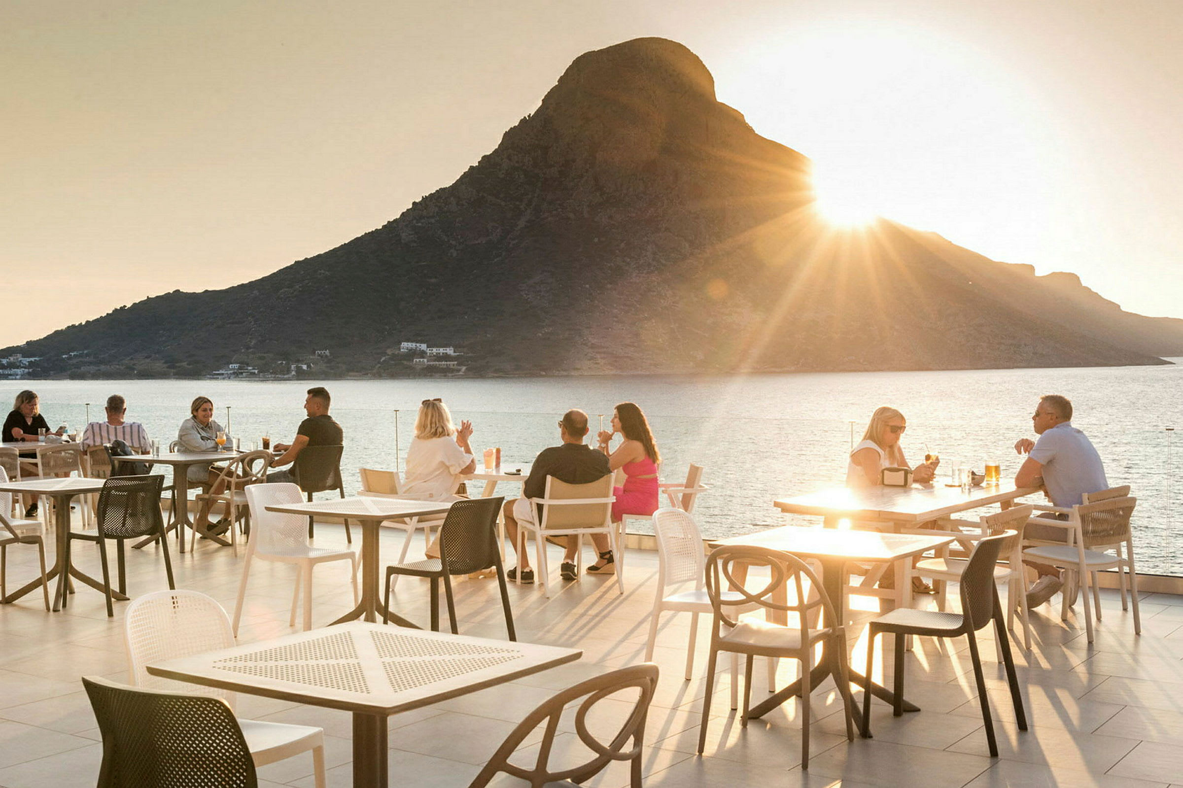 En utomhusterrass med sittplatser och ett berg vid havet i bakgrunden vid solnedgång. (AI-genererad text)