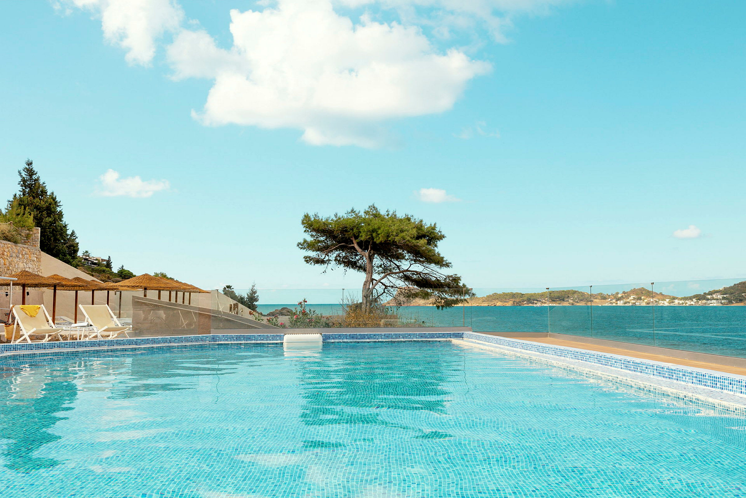Pool med blått vatten, omgiven av solstolar och paraplyer, med havsutsikt och en ensam träd i bakgrunden. (AI-genererad text)
