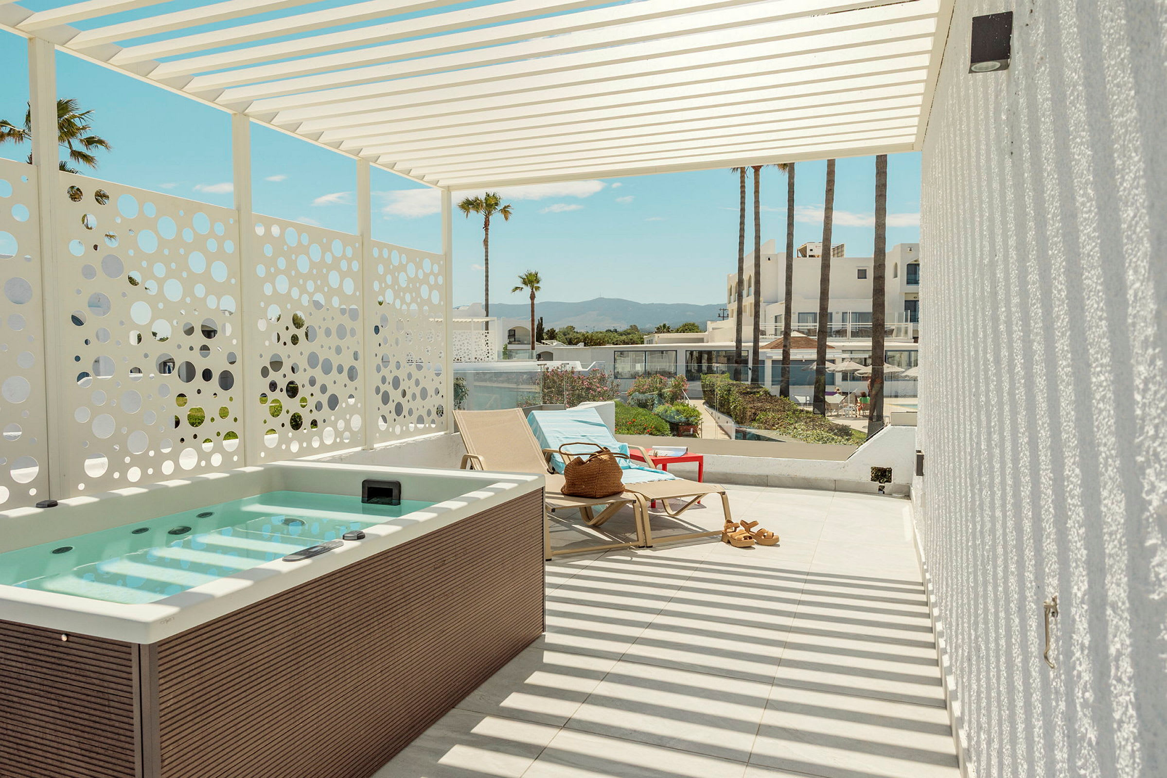 En terrass med jacuzzi, solstolar och utsikt mot palmer och byggnader i bakgrunden. (AI-genererad text)
