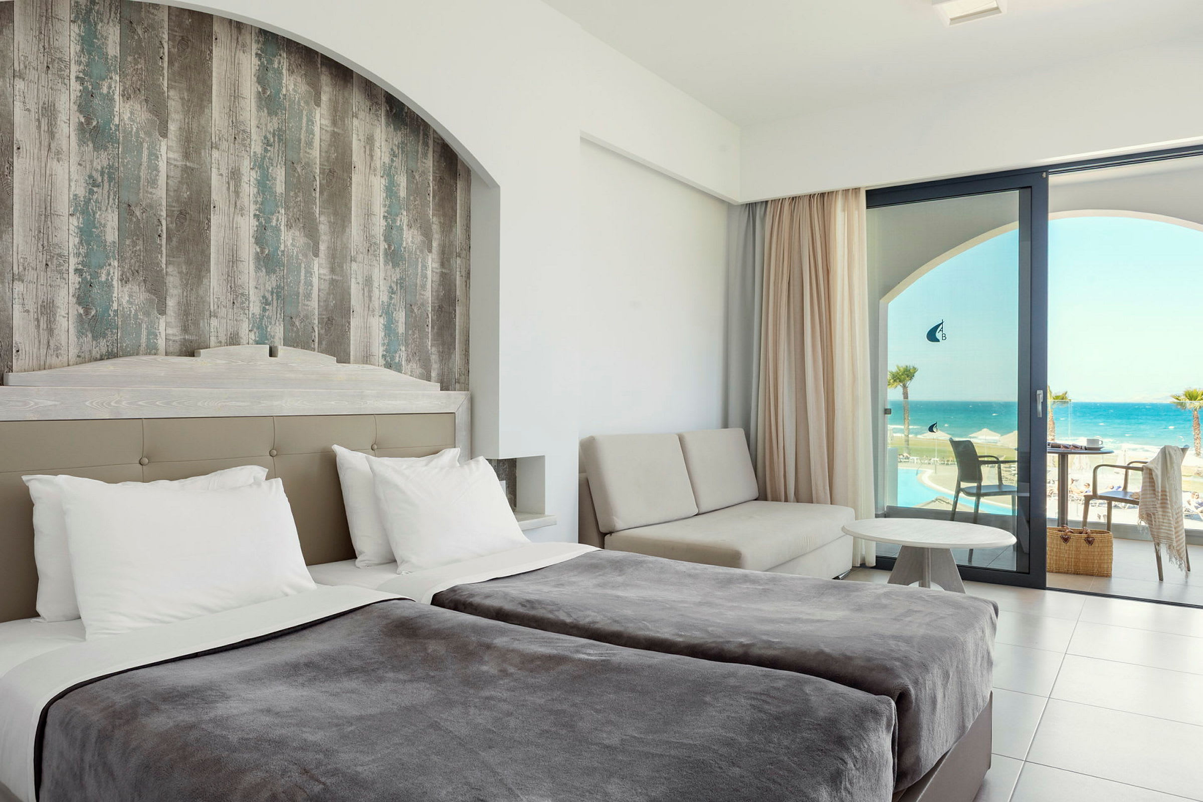 Hotellrum med dubbelsängar, en sittgrupp och utsikt över havet genom en glasdörr. (AI-genererad text)