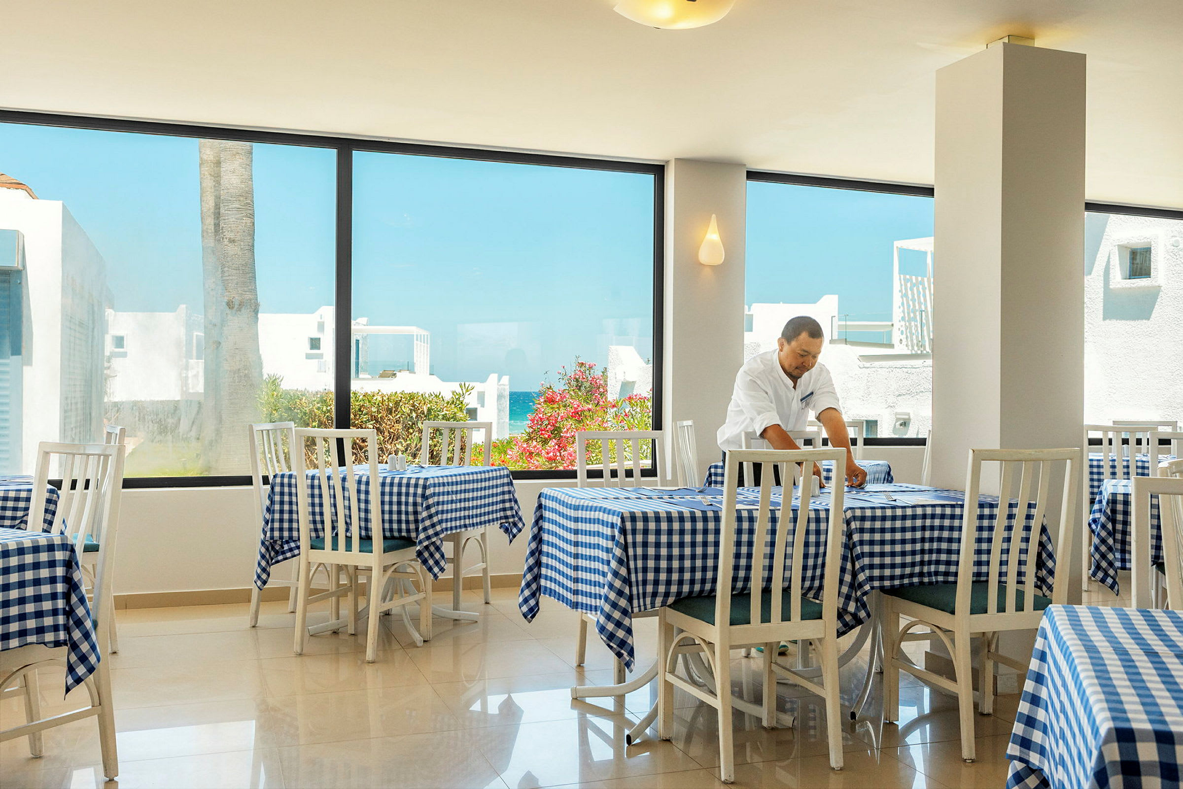 Ljus restaurang med vita stolar och rutiga blå dukar, samt stora fönster som visar utsikt över byggnader och grönska. (AI-genererad text)