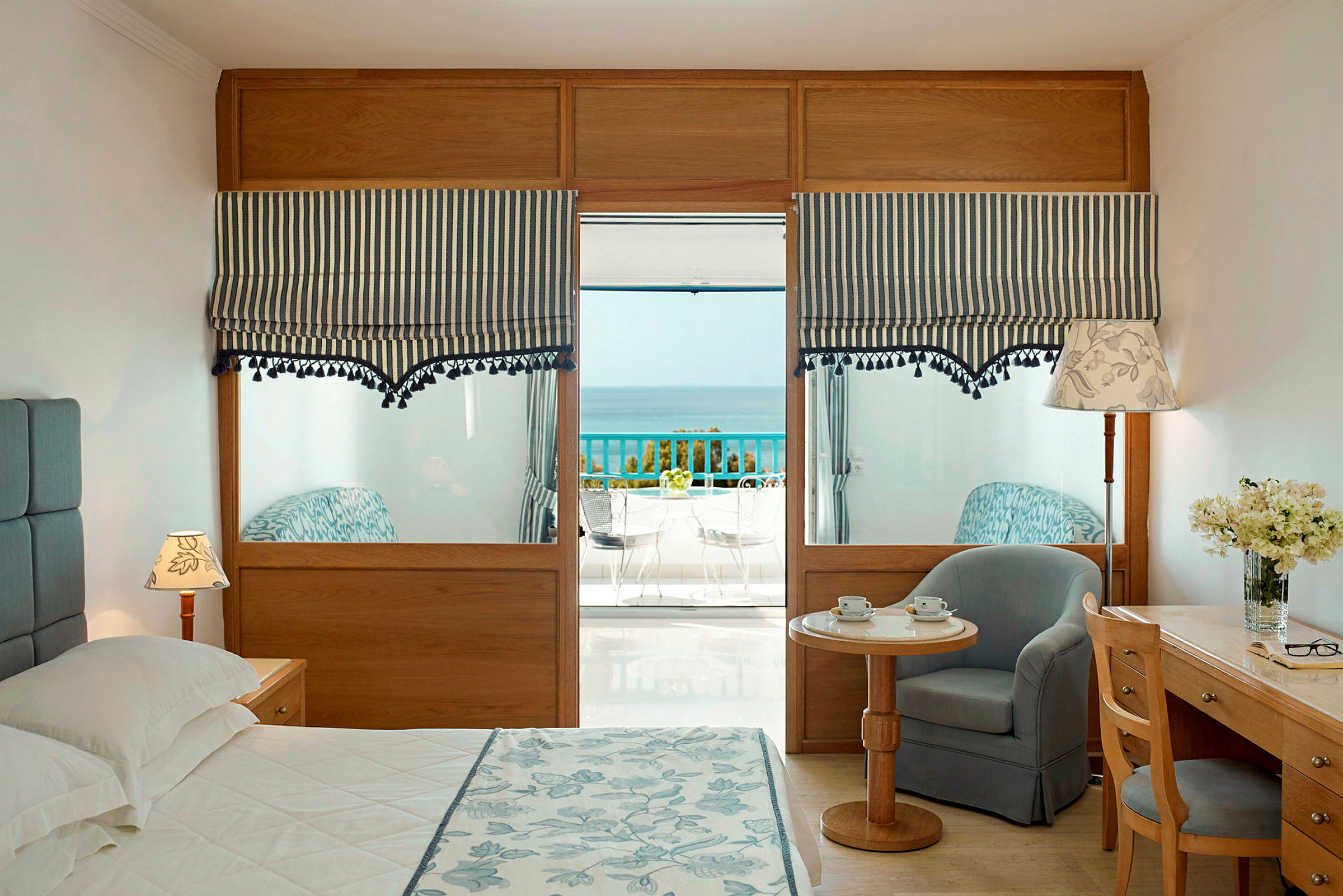 Ett hotellrum med en säng, en sittgrupp, ett skrivbord och utsikt mot havet genom fönster med randiga gardiner. (AI-genererad text)