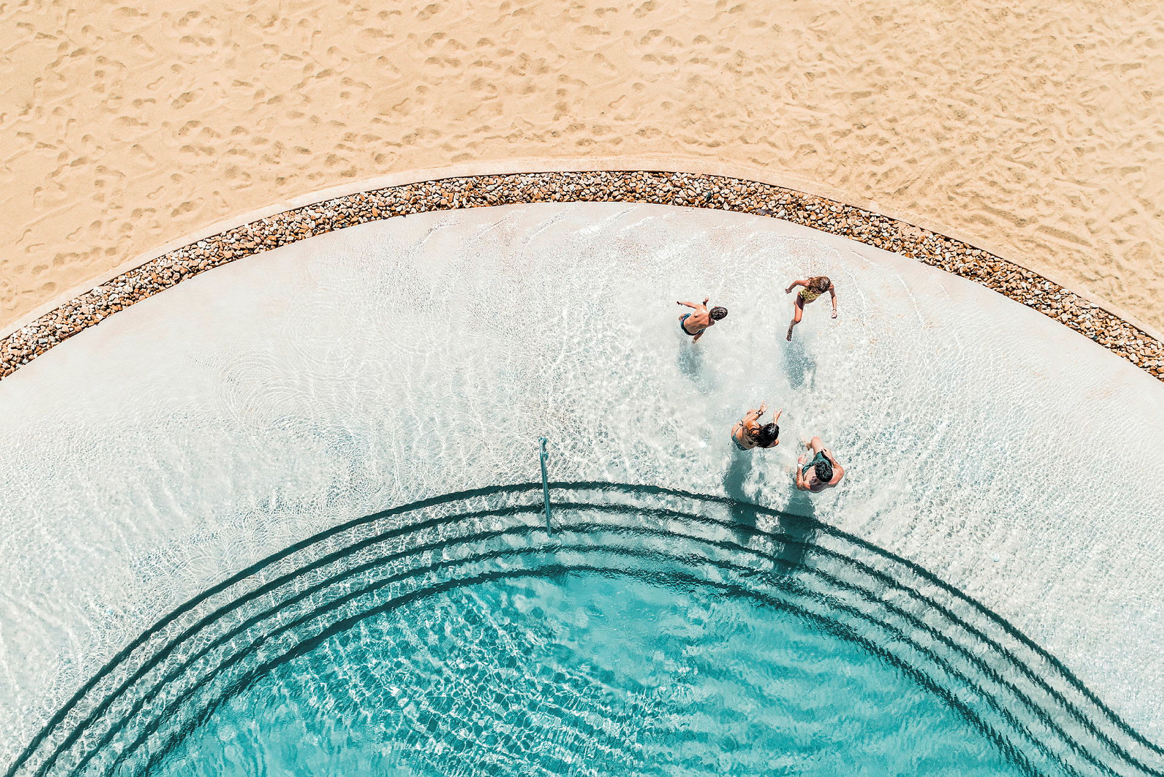Vy ovanifrån av en swimmingpool med människor som badar nära en strand med sand. (AI-genererad text)