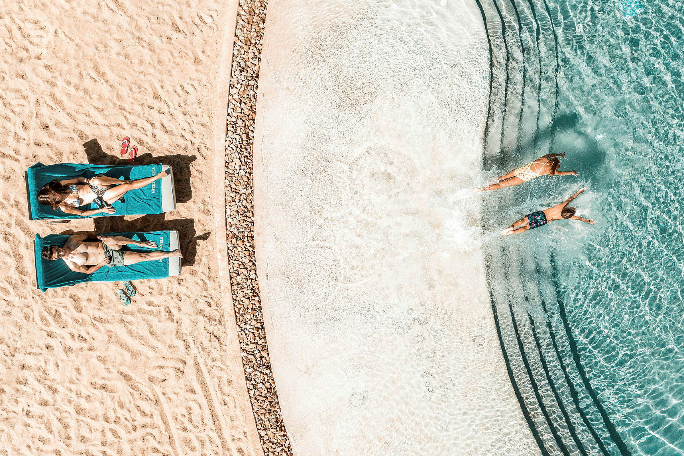 Flygfoto av två personer som ligger på solstolar på en strand bredvid en klarblå pool där två andra personer simmar. (AI-genererad text)
